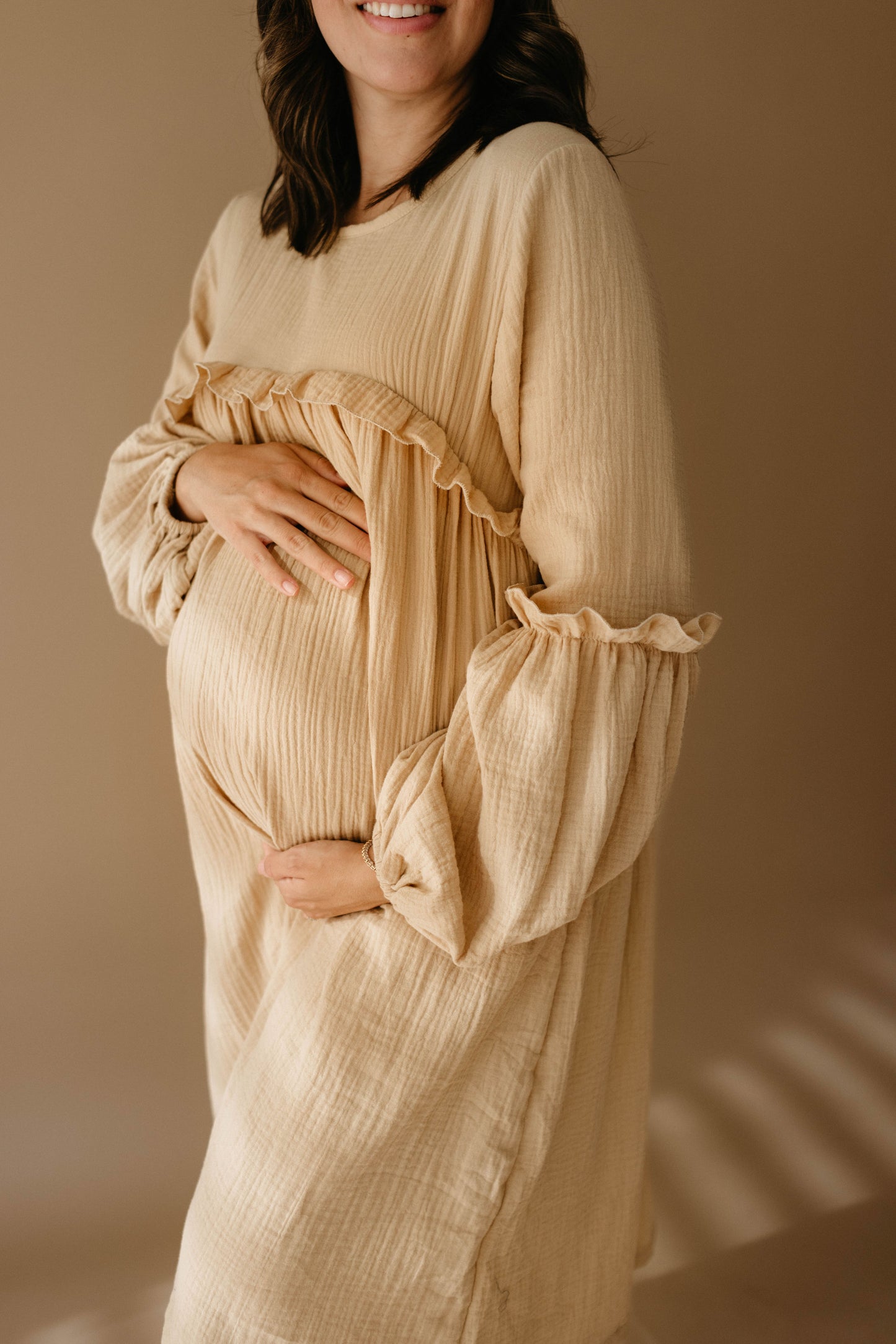 A pregnant woman wearing the "Golden Hour" long-sleeved dress by forever french baby gently cradles her belly with both hands. The neutral background accentuates the warmth and softness of the scene, evoking thoughts of matching rompers and child-size dresses to come.