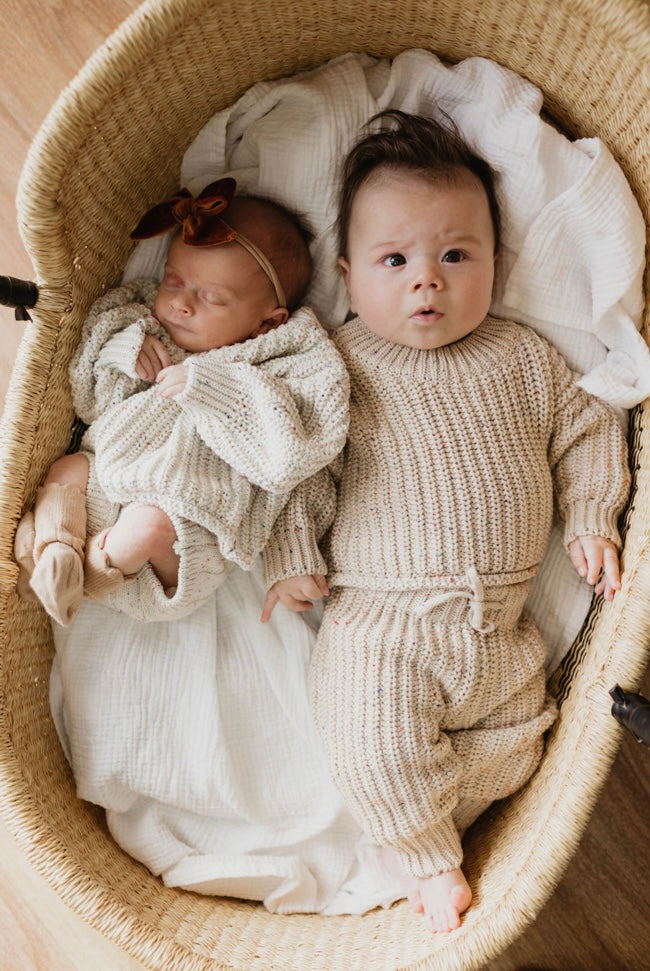 Two babies lie in a wicker bassinet, dressed in cozy knit sweaters. The baby on the left has a headband with a bow and is sleeping. The baby on the right has an expressive face and eyes open, wearing the Knit Sweater in Wheat Confetti by forever french baby along with matching pants. Both babies showcase an adorable baby wardrobe as they rest on a white blanket.