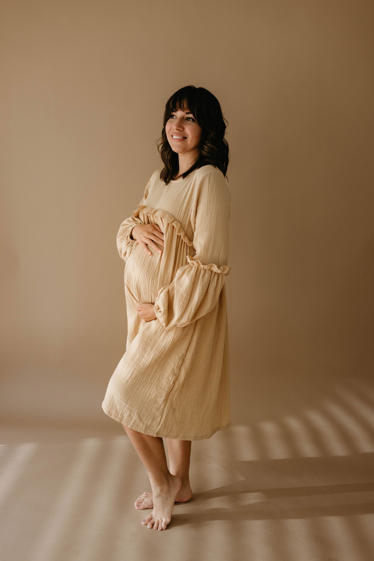 A pregnant woman in a "Golden Hour" dress by forever french baby stands barefoot against a neutral background, smiling and holding her belly with both hands, her thoughts perhaps drifting to matching rompers for the little one on the way.