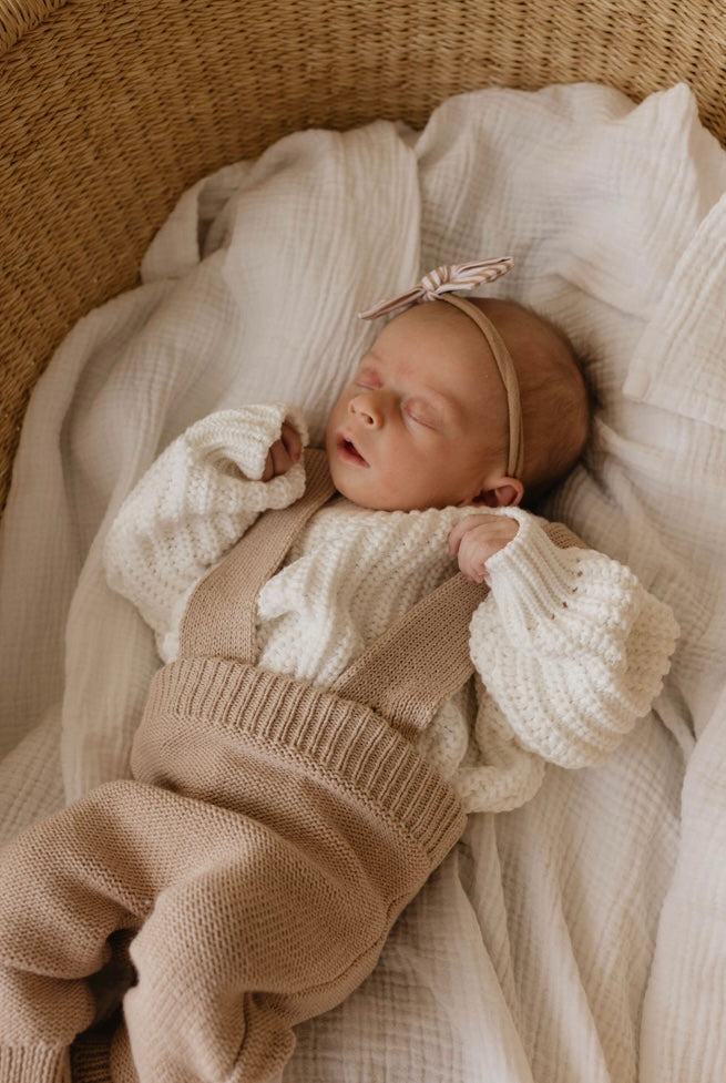 A newborn baby peacefully sleeps in a wicker basket lined with a white blanket. The baby is dressed in the Forever French Knit Suspenders | Graham, paired with a white sweater and a beige headband featuring a small bow. The natural light illuminates the cozy and warm setting, perfect for cooler weather.