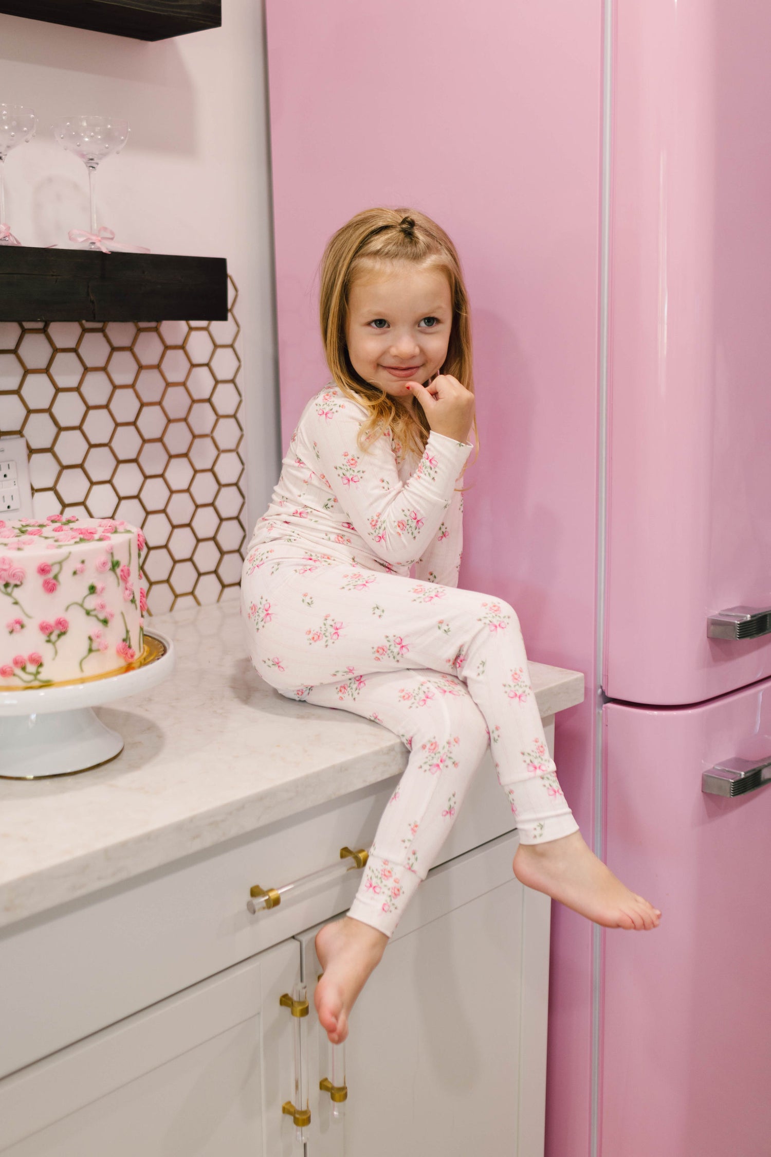 A young girl in Kendy 2's Bamboo Two Piece Pajamas | Kendy Floral sits on a kitchen counter beside a pink refrigerator. She smiles with her hand on her chin. Below the counter is a cake decorated with floral patterns. The soft bamboo fabric and adorable pink bows add charm to the modern kitchen with its hexagonal tile backsplash.