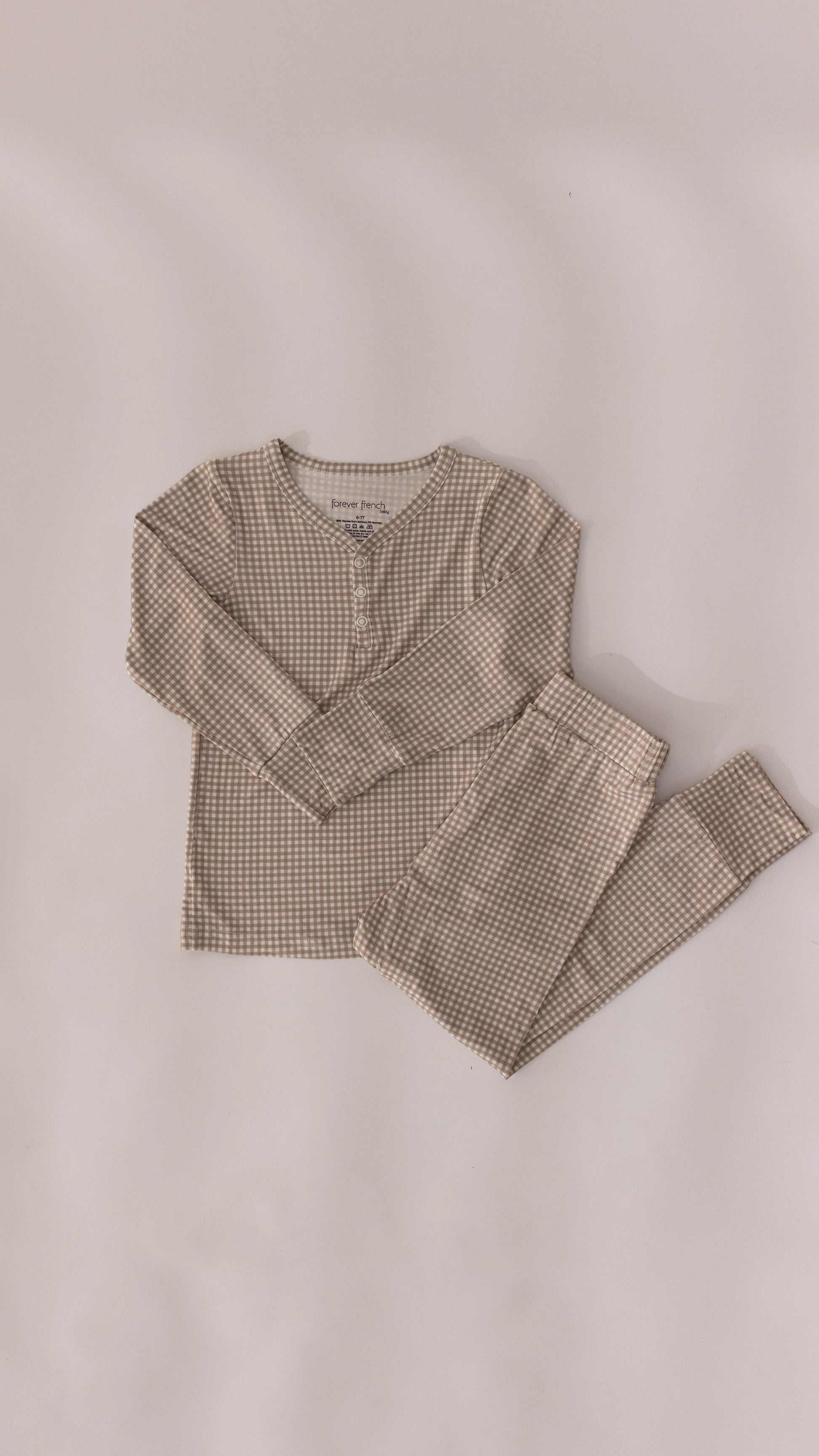 Introducing the Ryan Gingham Bamboo Two Piece Pajamas from forever french baby—a delightful set of children's pajamas featuring a long-sleeved top and matching pants. This hypo-allergenic ensemble showcases a charming beige and brown checkered pattern, laid out flat on a light surface. The top is adorned with buttons at the neckline, providing both style and comfort for your little one.