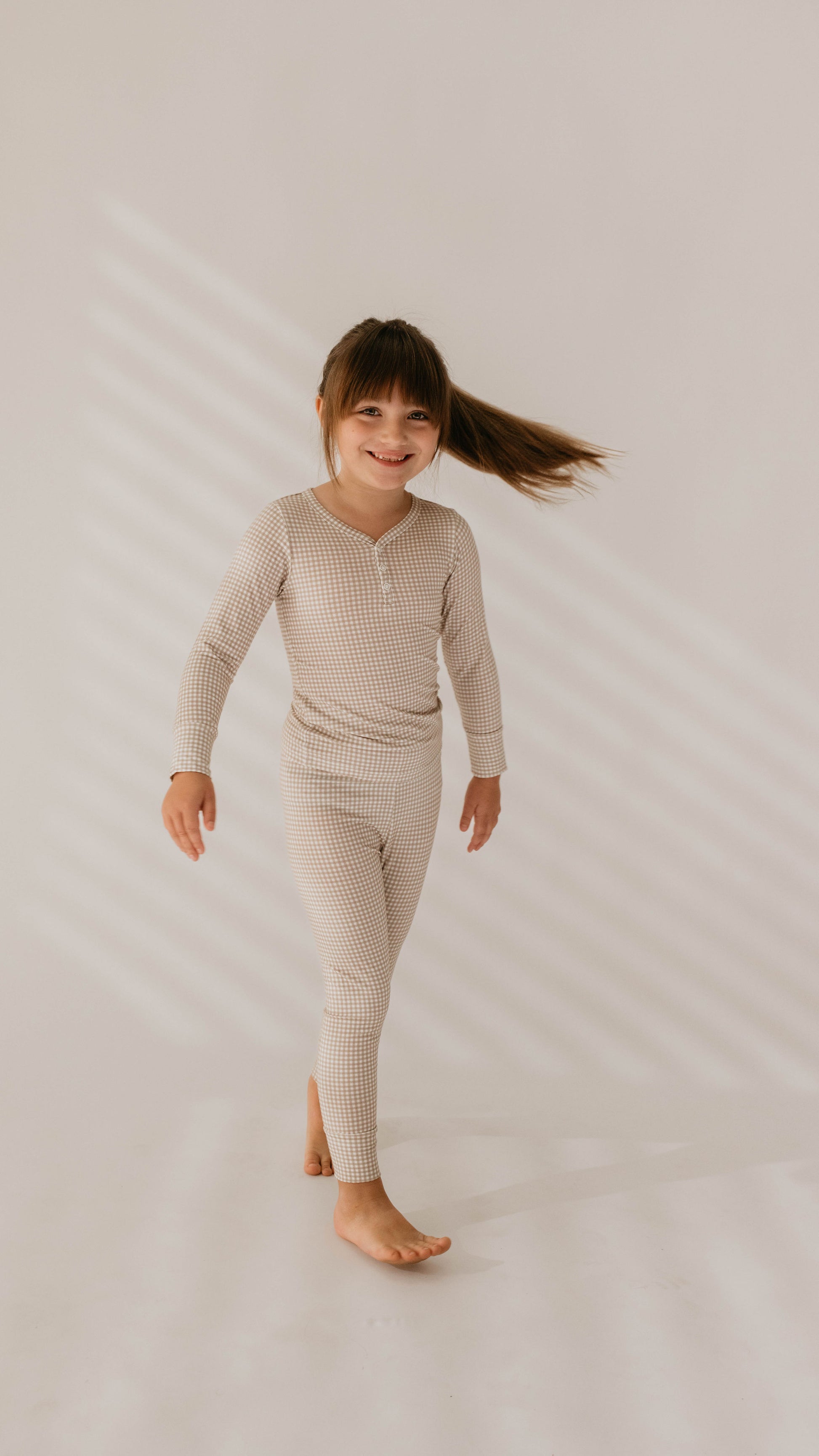 A young child with long brown hair in a ponytail is smiling and walking barefoot. They are wearing the Ryan Gingham Bamboo Two Piece Pajamas from forever french baby, featuring a neutral-colored checkered design made from hypo-allergenic fabric. The background is minimal with a soft stripe shadow pattern on the wall.