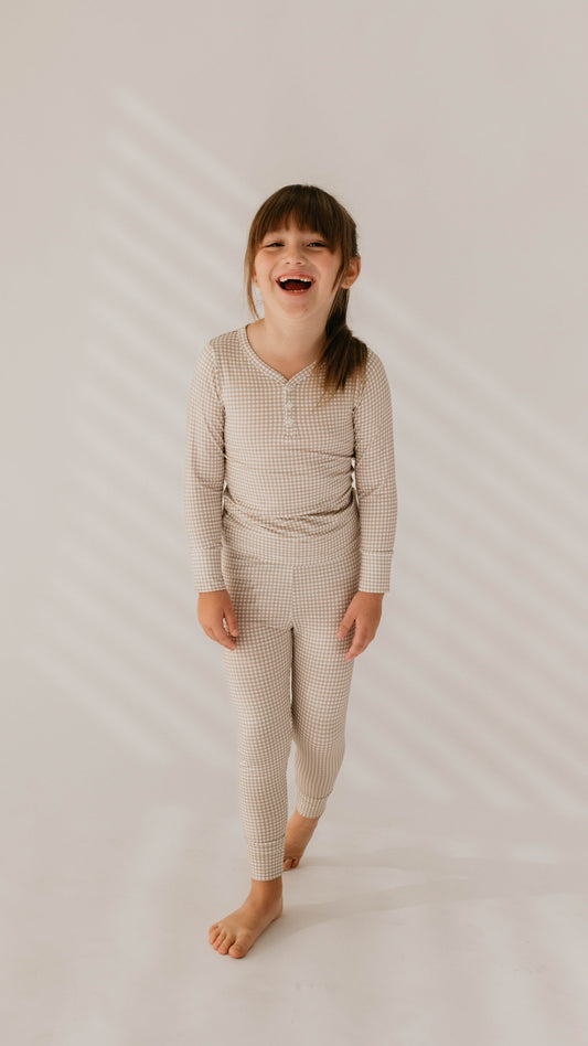 A young child with long hair, smiling widely and standing in a relaxed pose, is wearing a matching set of light-colored, checkered "Ryan Gingham" Bamboo Two Piece Pajamas from forever french baby and is barefoot. The background features plain walls with soft, slanted light patterns that create a cozy atmosphere.