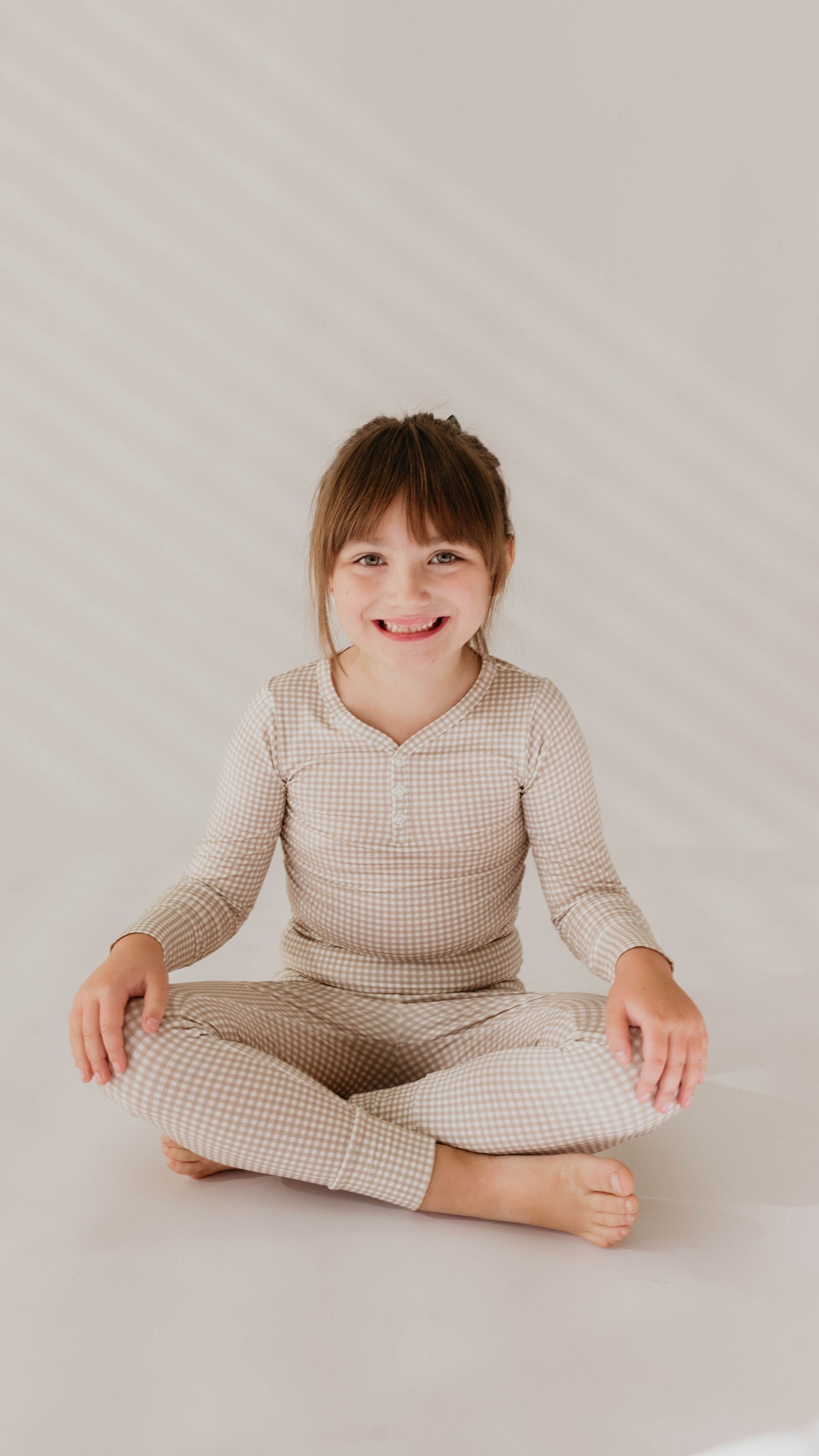 A young child with brown hair and bangs is sitting cross-legged on the floor, smiling at the camera. They are wearing the Ryan Gingham Bamboo Two Piece Pajamas from forever french baby against a plain, light-colored background.