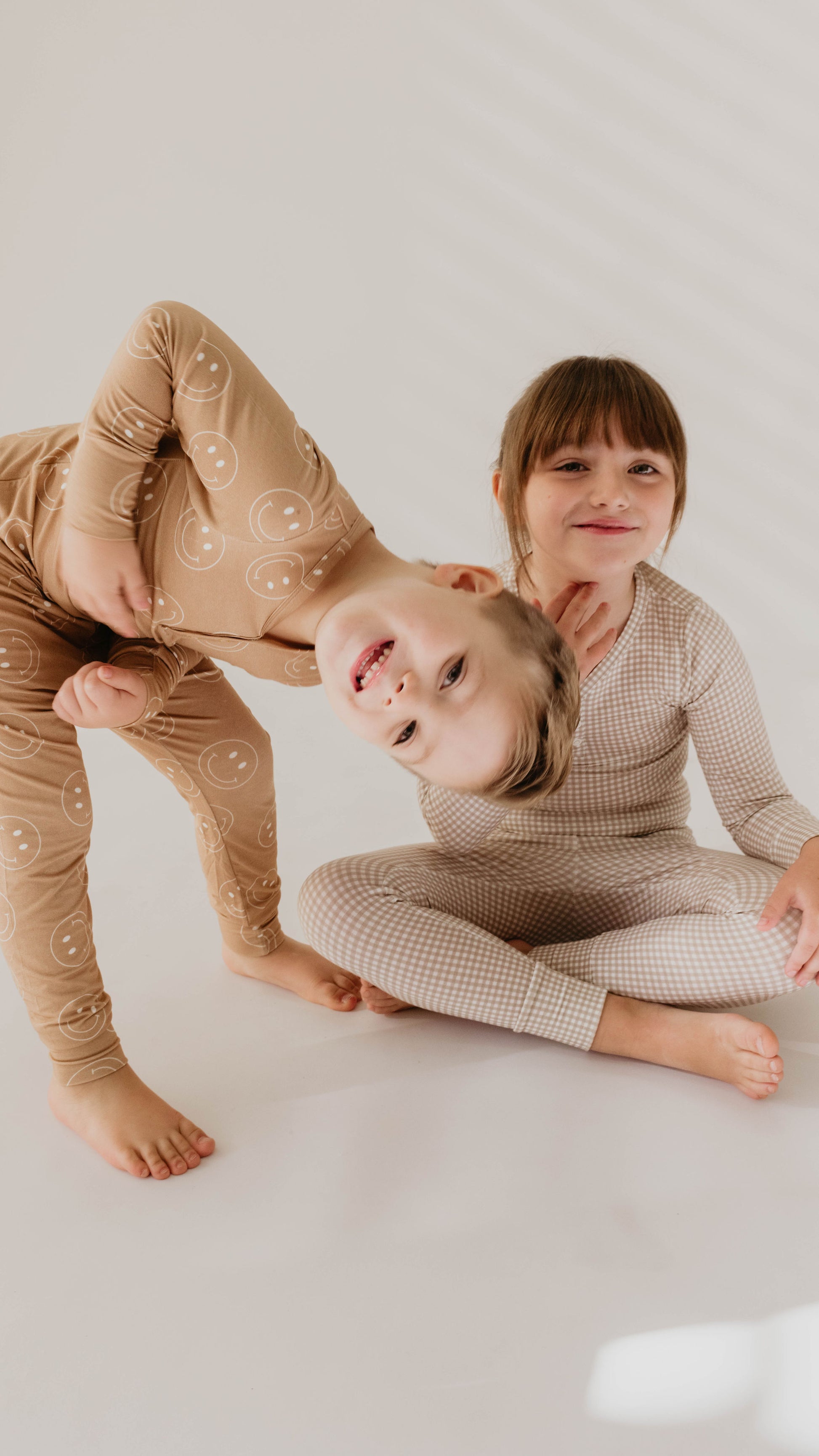 Two children in matching Ryan Gingham bamboo pajamas from forever french baby. One child is seated cross-legged, smiling at the camera, while the other stands bent over at the waist, leaning sideways and smiling upside down. Both are barefoot and appear playful and happy in their hypo-allergenic clothing.