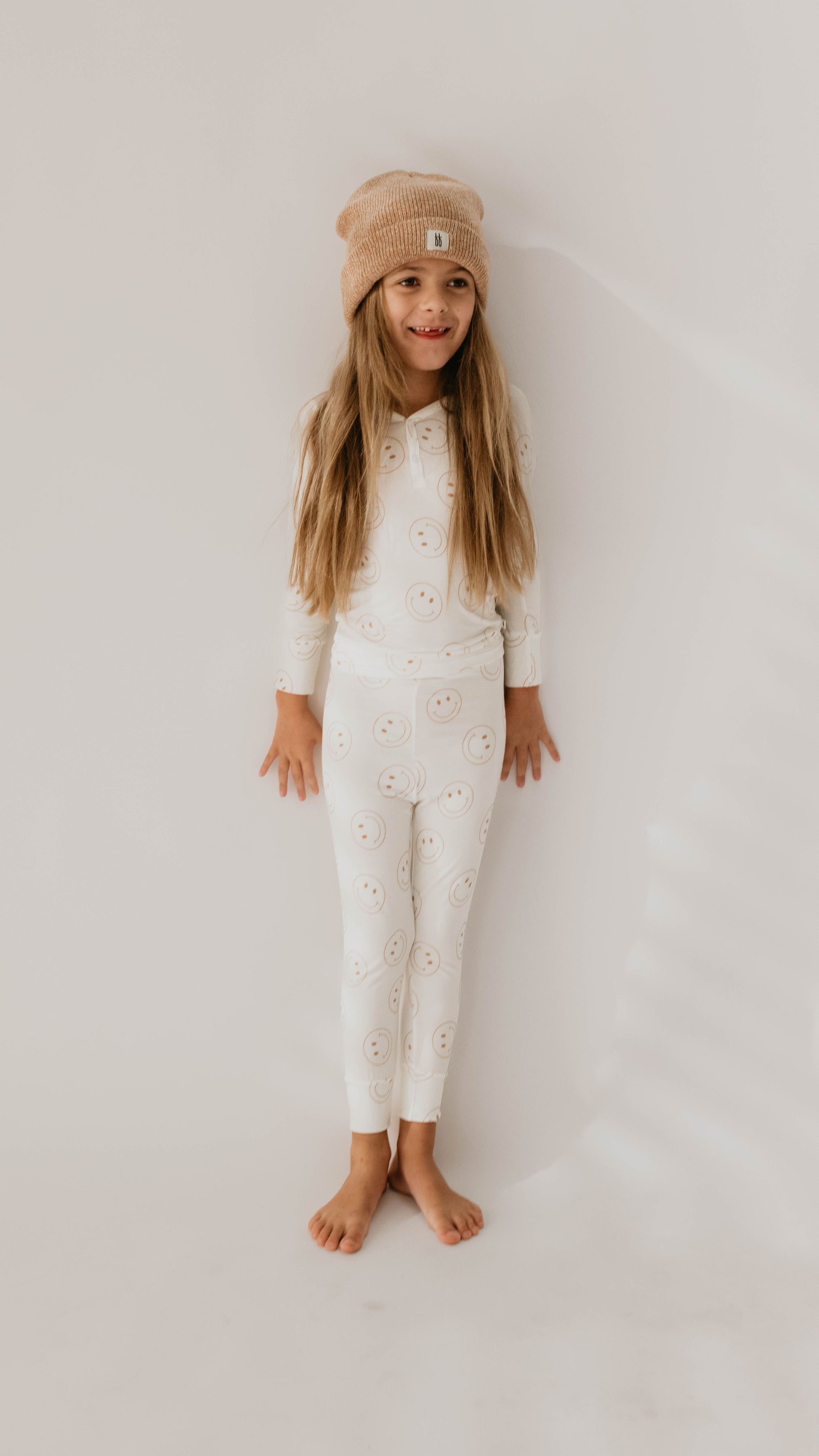 A young child with long hair stands against a plain white background, barefoot and smiling. They are wearing a matching "Bamboo Two Piece Pajamas | Just Smile" from forever french baby, adorned with a smiley face pattern, and a brown knit beanie.