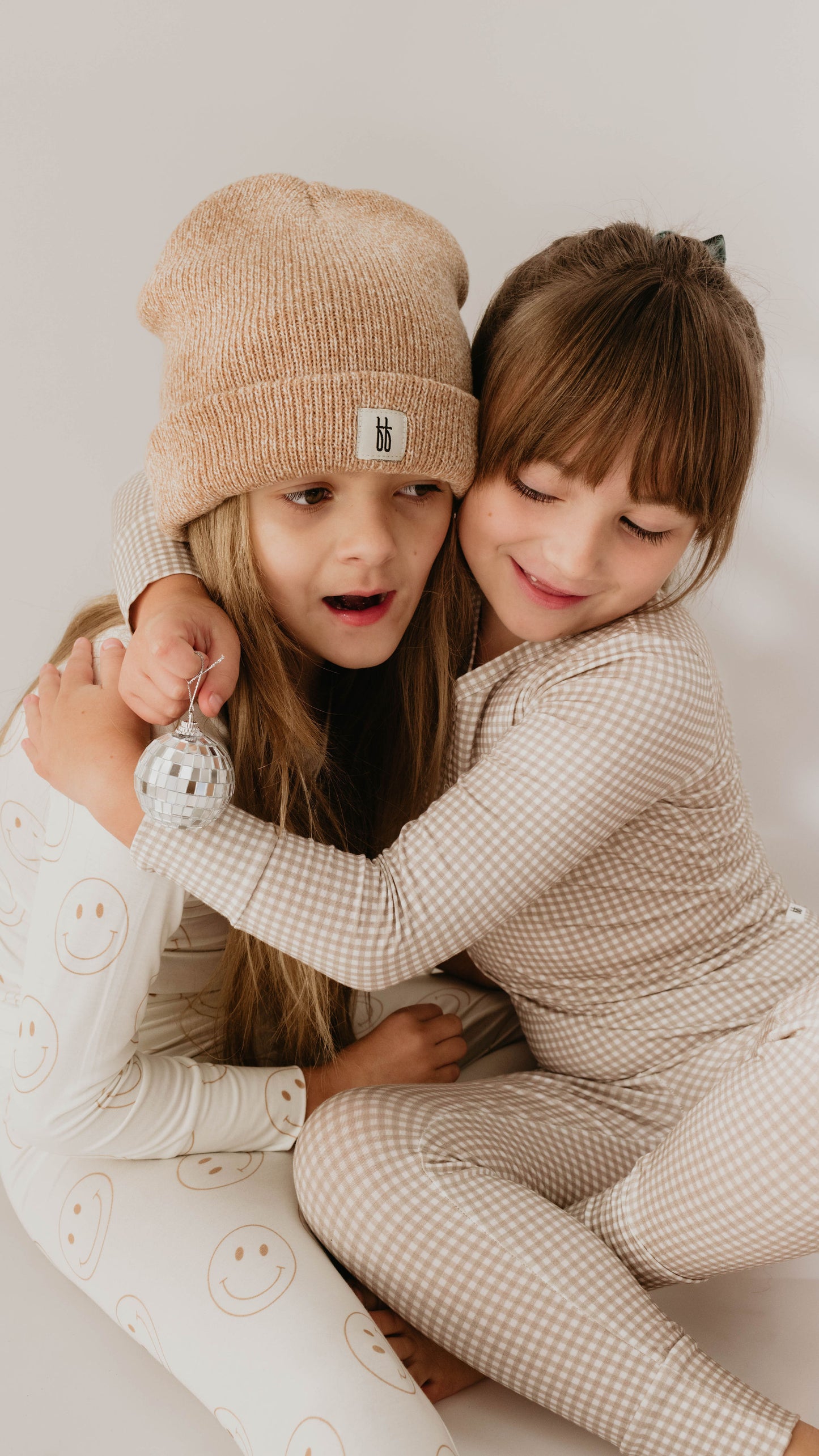 Two children, dressed in cozy, patterned bamboo pajamas from forever french baby, sit close together against a neutral background. One child, wearing a knitted beanie and smiling face pajamas, holds a small silver ornament. The other child, in Ryan Gingham hypo-allergenic Bamboo Two Piece Pajamas from forever french baby, hugs the first child warmly.