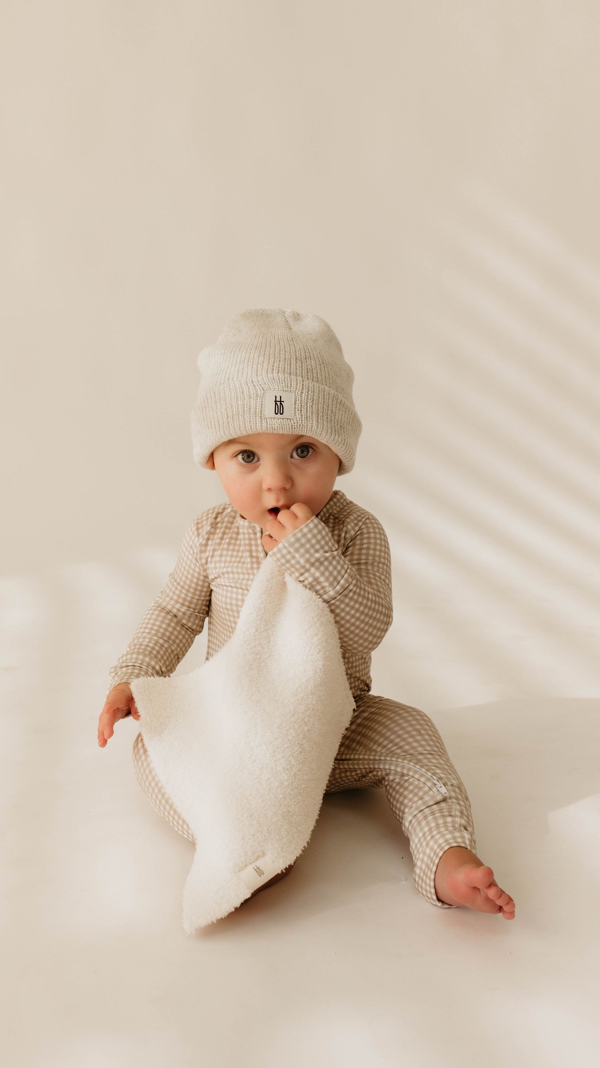 A baby, dressed in a checkered onesie and a knitted hat from forever french baby, sits on a light surface holding the Lovey | Cream, looking directly at the camera with wide eyes. Soft shadows from window blinds are cast in the background.