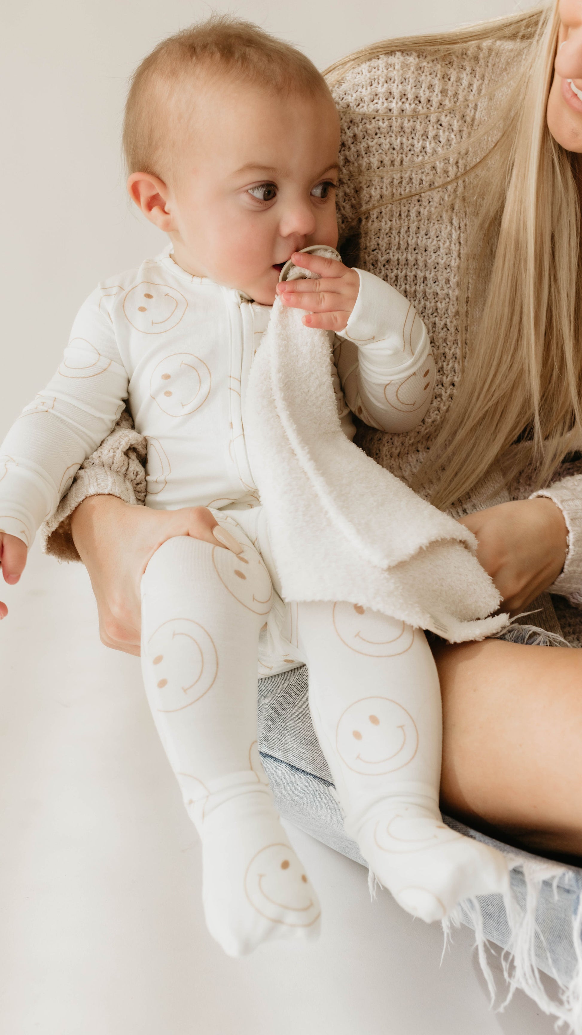 A baby in white pajamas with smiley faces holds a Lovey | Cream from forever french baby and touches their mouth while sitting on a woman's lap. The woman, with long blonde hair and wearing a luxurious beige microfiber feather yarn sweater, is partially visible, holding the baby securely. The background is neutral.