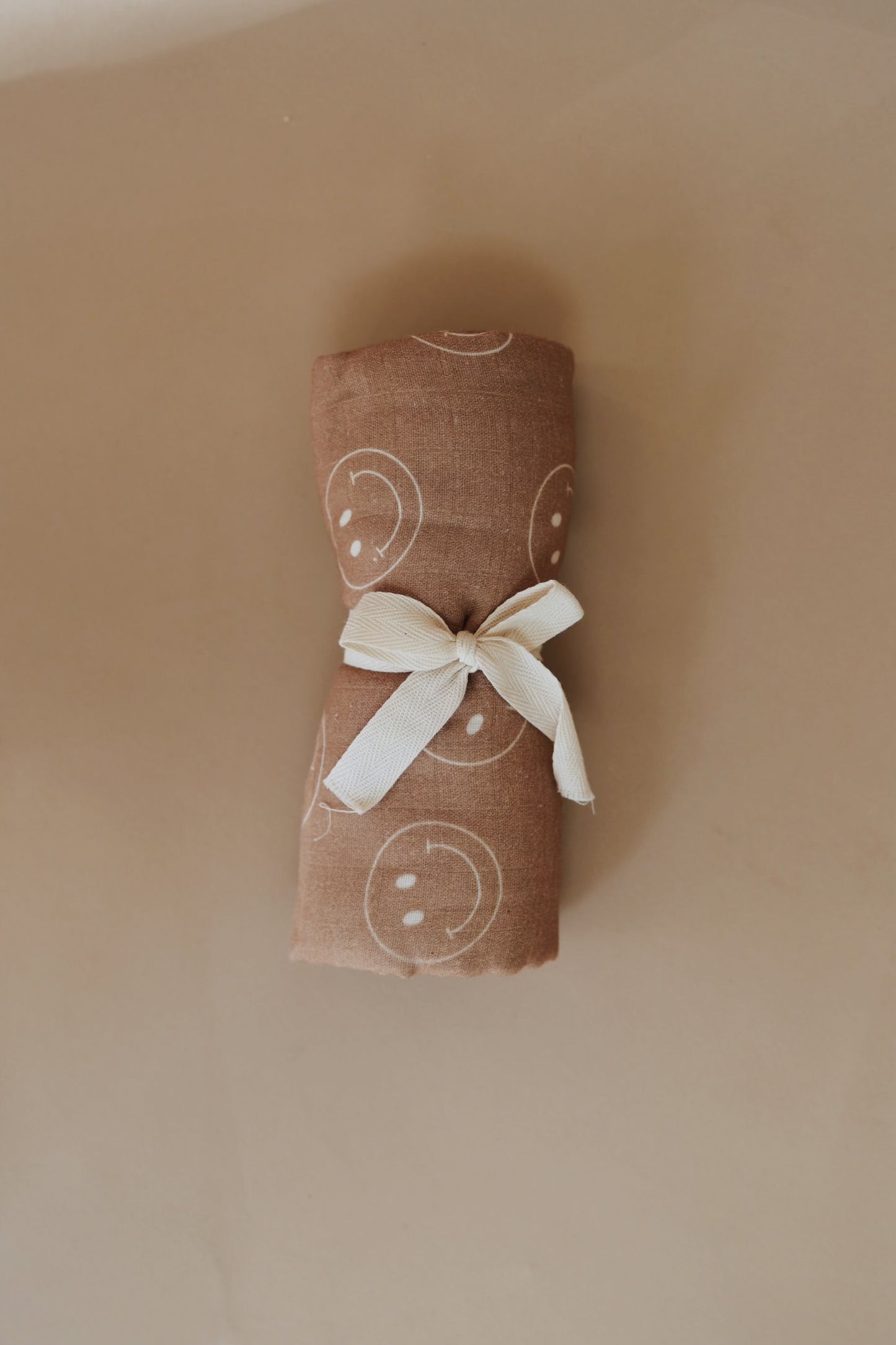 A neatly rolled Muslin Swaddle | Just Smile Tan with white smiley face prints is tied with a white ribbon on a beige background. The forever french baby fabric roll is centrally positioned, and the ribbon forms a simple bow.