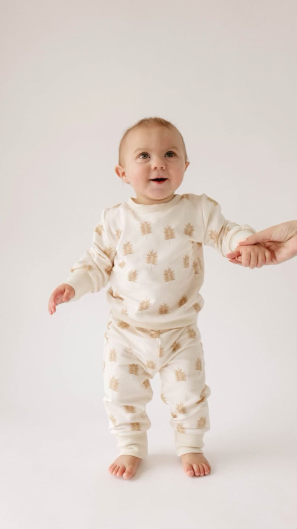 A smiling baby in a beige and white Palm Sweater Set from forever french baby, featuring a sun pattern, stands holding an adult's hand against a plain white backdrop.