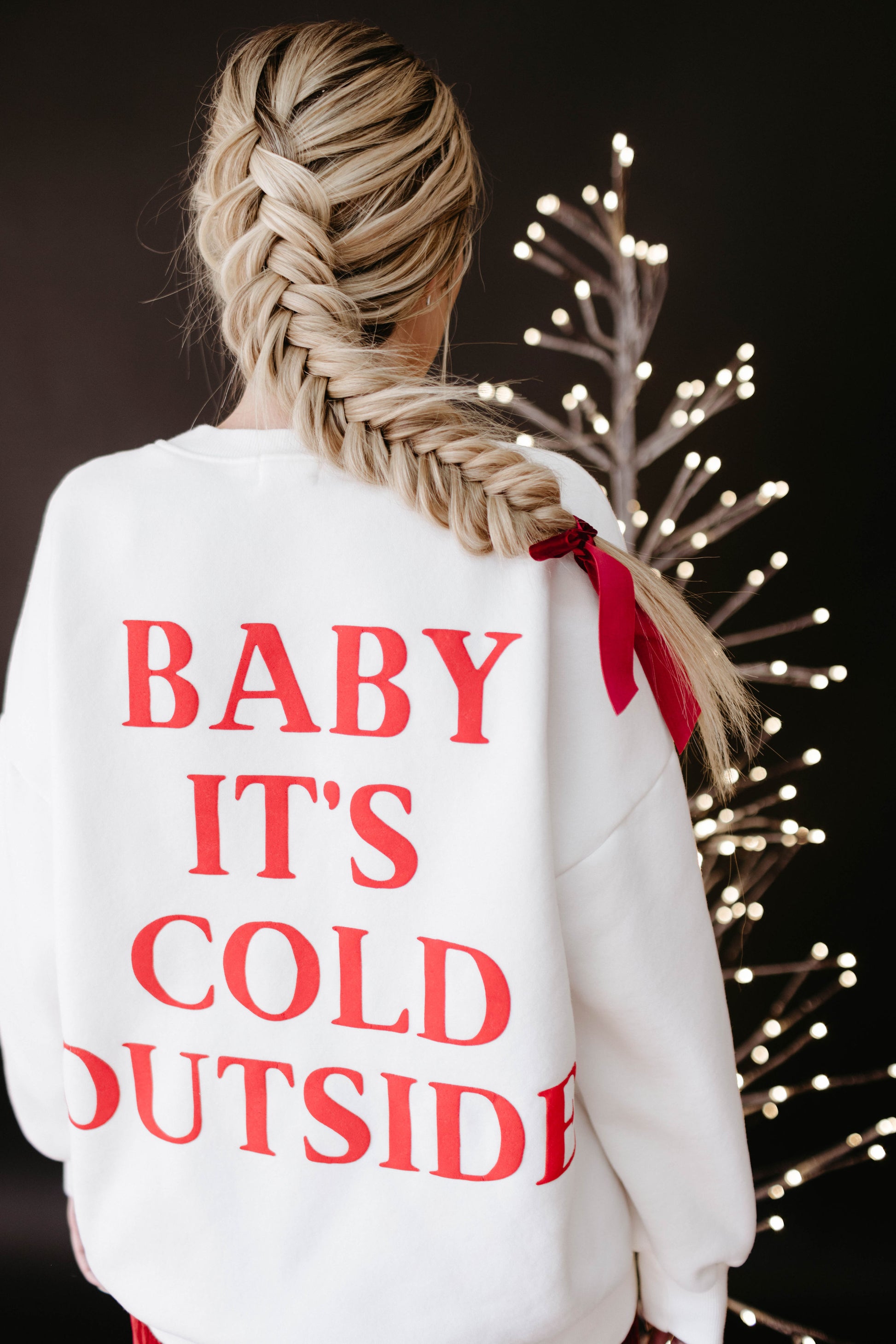A person with a long braided hairstyle, accentuated by a red ribbon, is wearing an "Adult Sweatshirt" from forever french baby. The sweatshirt features the phrase "BABY IT'S COLD OUTSIDE" in red text on a white background. They are standing in front of a lit decorative tree, which perfectly captures the warmth of the holiday season against the dark backdrop.