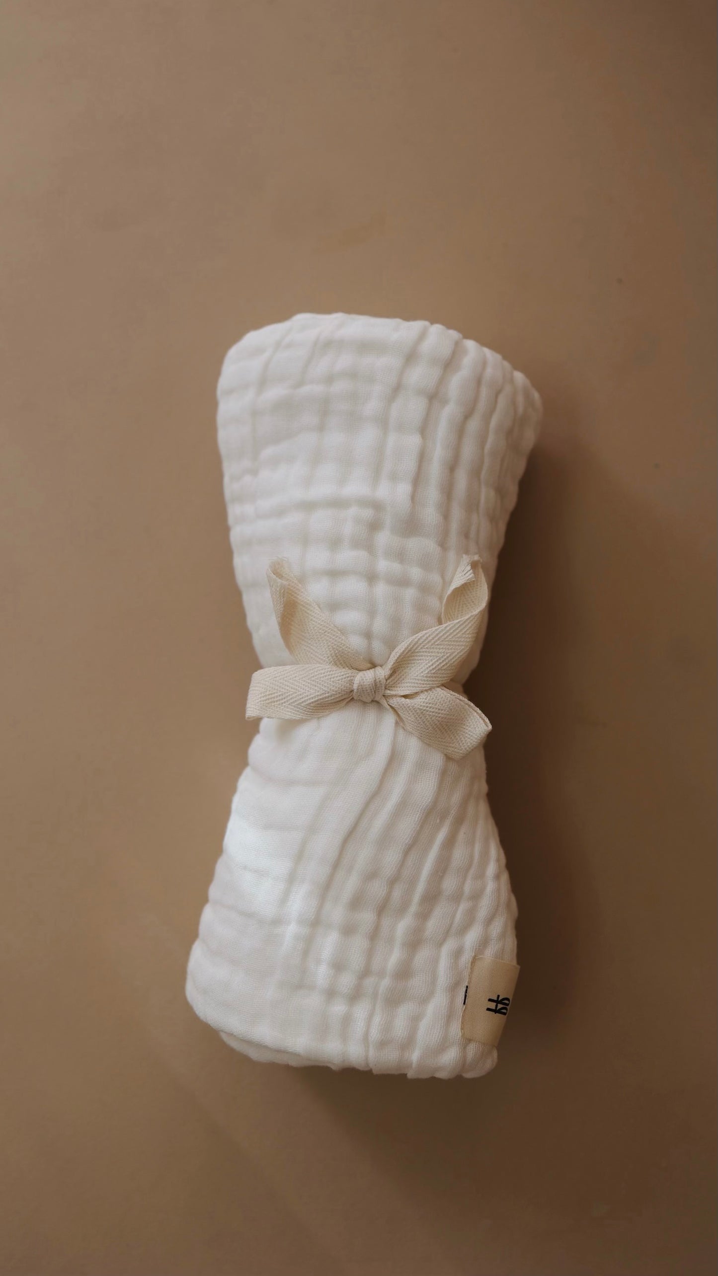 A rolled-up, white, textured crib sheet from Forever French Baby is tied with a beige ribbon and placed on a light brown background. The sheet features a subtle, quilted pattern and has a small tag on one edge reading "Forever French Baby." For the best care, wash with like colors and tumble dry on low.