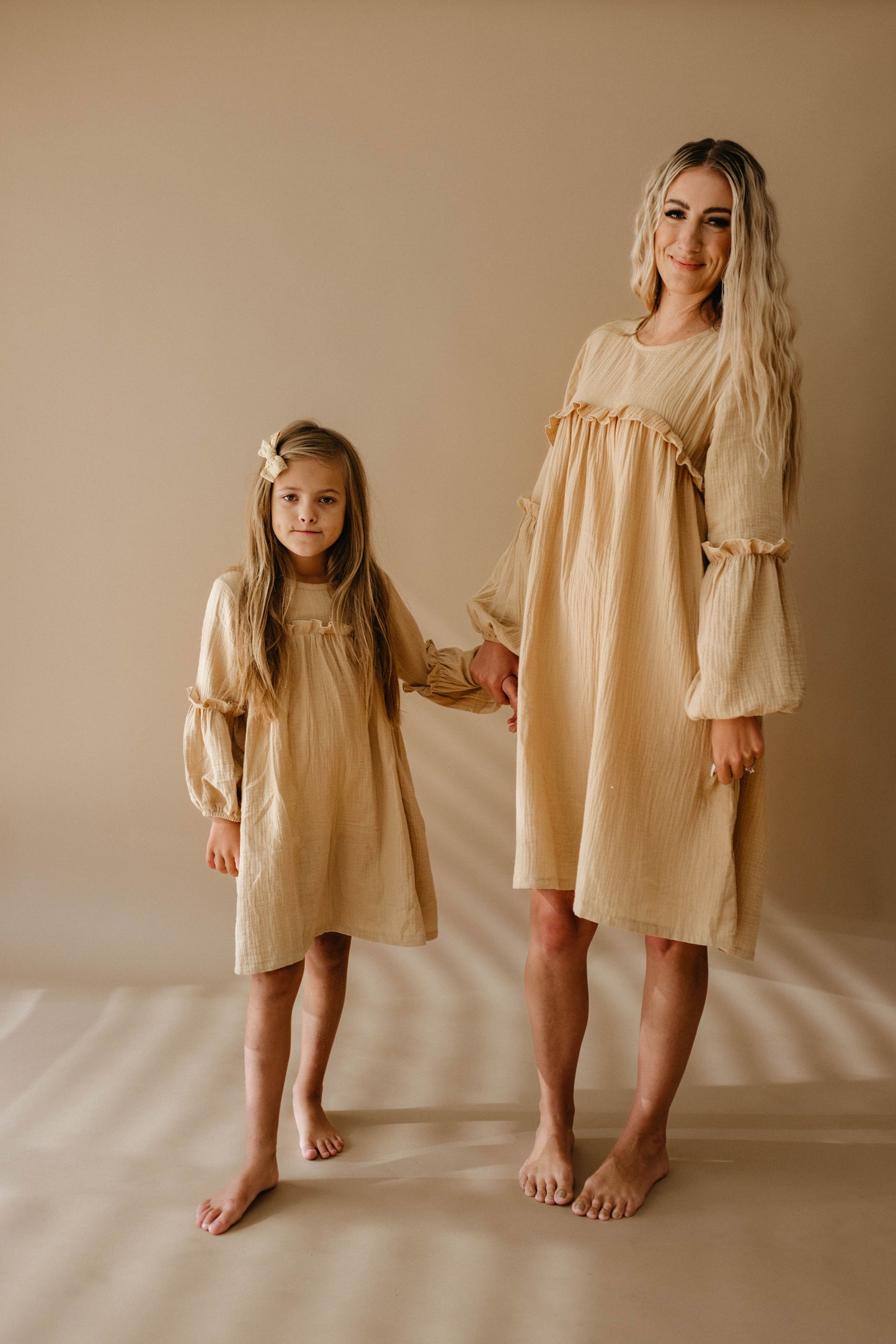 A woman with long, wavy hair and a young girl with a bow in her long hair stand barefoot on a beige background, holding hands and wearing matching dresses from the "Women's Dress | Golden Hour" collection by forever french baby. They are smiling softly.