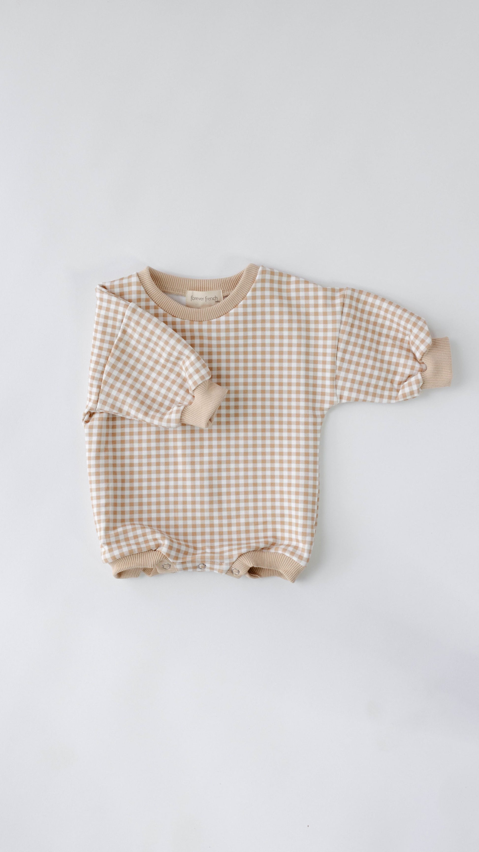 The Goldie Gingham | Sweatshirt Romper from forever french baby is a long-sleeved baby romper featuring a beige and white gingham pattern, complete with beige cuffs at the wrists, neck, and legs. Laid flat on an off-white surface, this romper adds charming elegance to any little one's wardrobe.