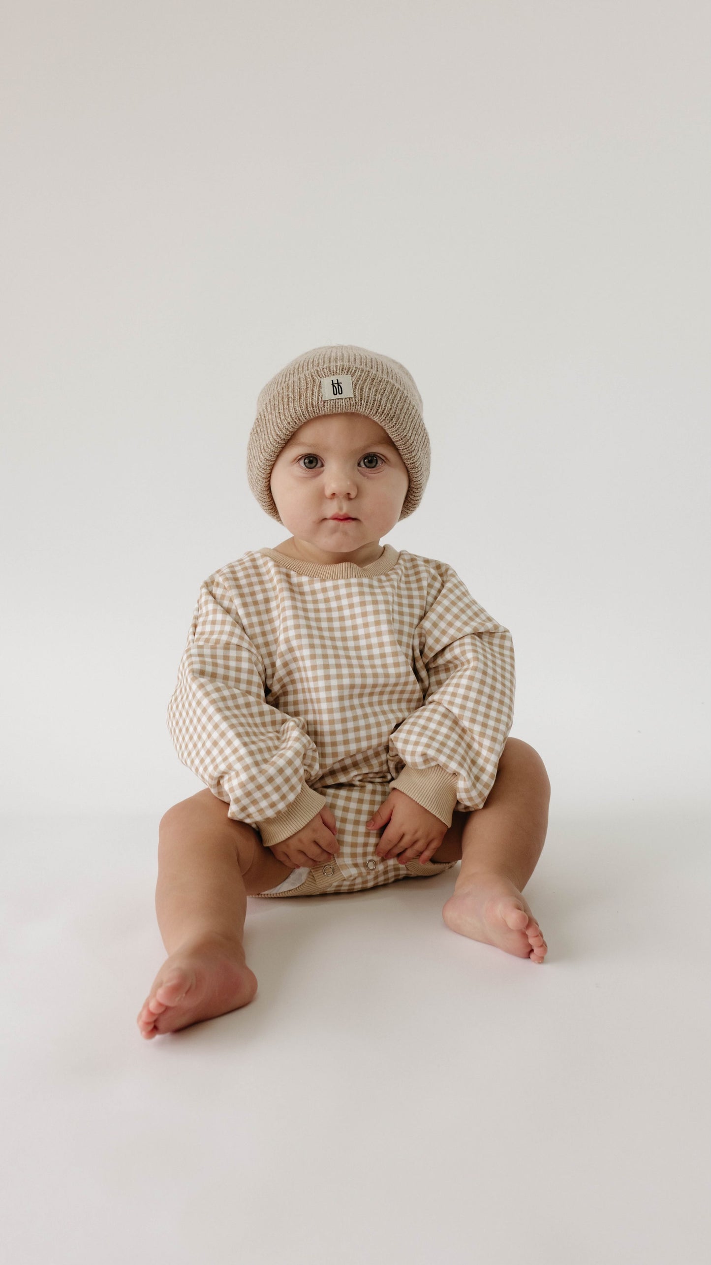 A toddler wearing the forever french baby Goldie Gingham | Sweatshirt Romper in a beige and white checkered pattern and a beige beanie sits on a white background with legs outstretched, looking ahead with a neutral expression.