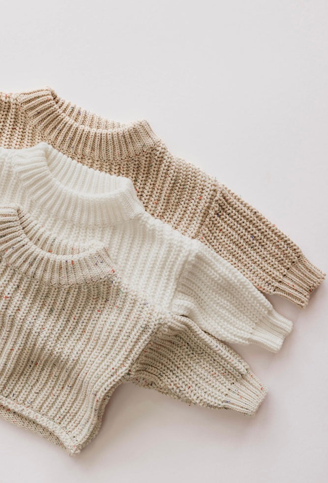 A top-down view of four "Knit Sweater | Wheat Confetti" sweaters by forever french baby in neutral colors stacked on a light surface. These classic knit sweaters feature a ribbed texture and high collars, with colors ranging from light beige to cream. Some showcase subtle variations of speckles or patterns—essential pieces for any baby wardrobe.