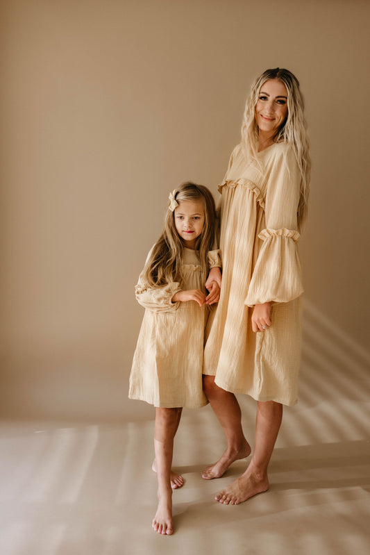 A smiling woman and a young girl stand barefoot in matching "Golden Hour" dresses by forever french baby, against a neutral background. The woman has long, wavy hair, and the girl has a bow in her hair. Both appear relaxed and happy.