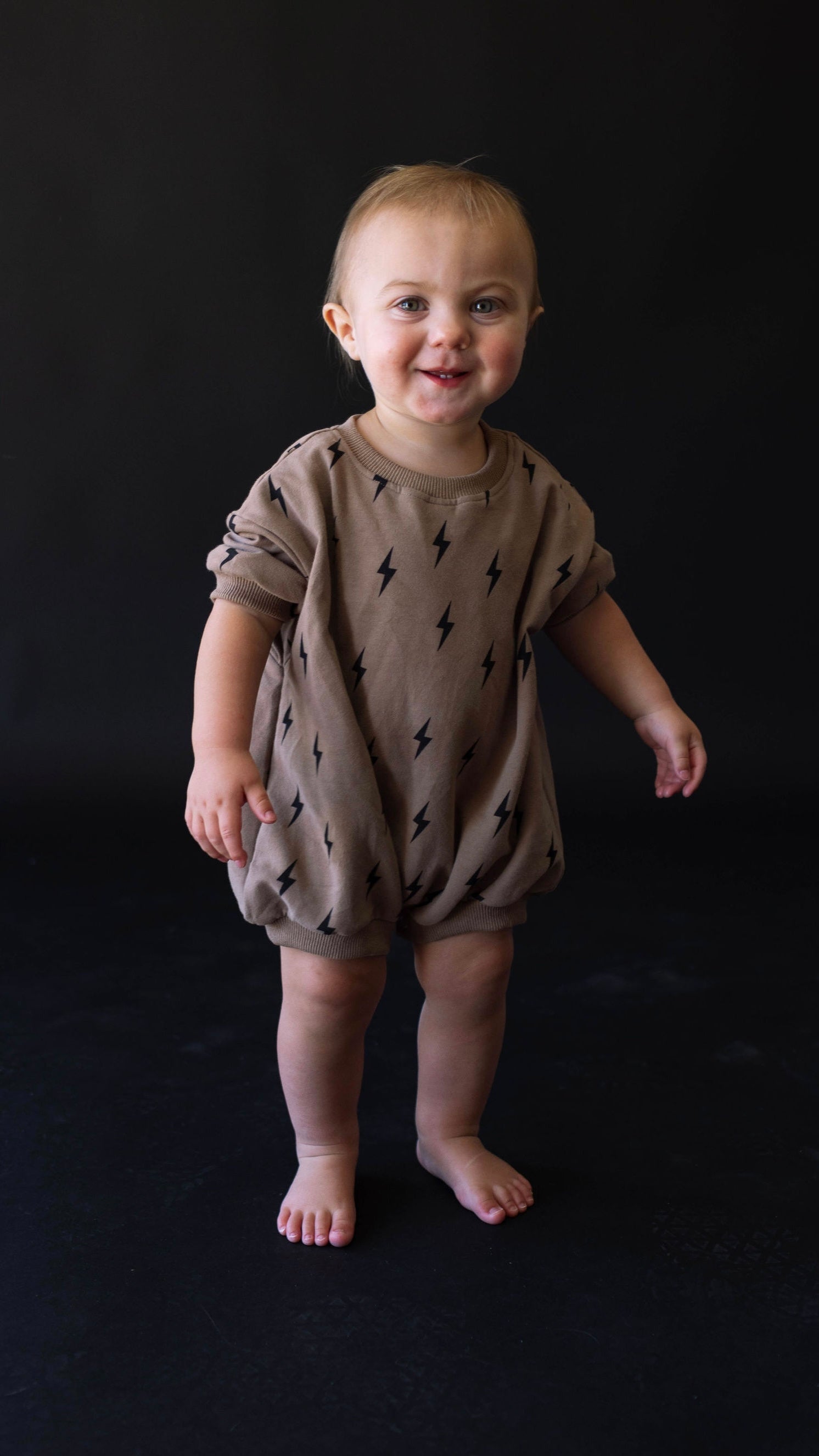 A toddler with short, blond hair stands barefoot against a dark background. The child is wearing a light brown Short Sleeve Romper | Lightning Bolt made of organic cotton by forever french baby. The child appears to be smiling and is looking directly at the camera.