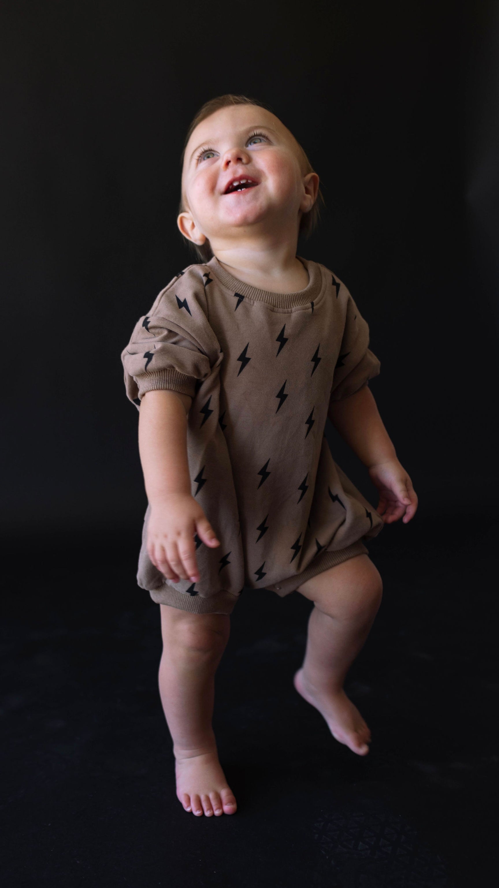 A toddler, dressed in a Short Sleeve Romper | Lightning Bolt by forever french baby, made of organic cotton, stands and smiles joyfully against a dark background. The child appears to be mid-motion, perhaps dancing or playing.