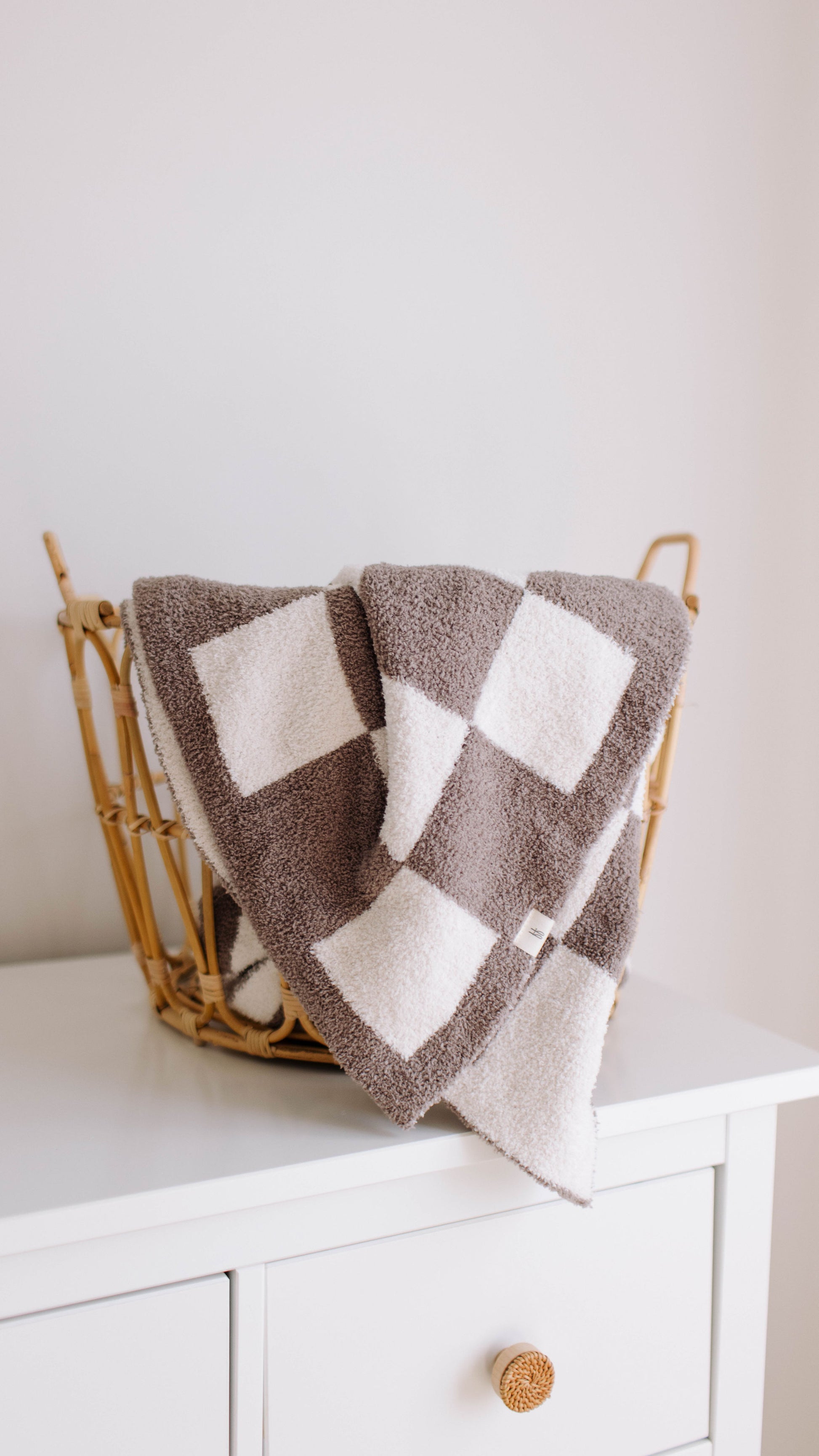 A Plush Blanket in Mushroom Checkerboard from forever french baby, with a velvety-rich texture, is draped over the edge of a wicker basket. The basket is placed on top of a white dresser with a single visible drawer that has a round, textured knob. The background is plain and light-colored.