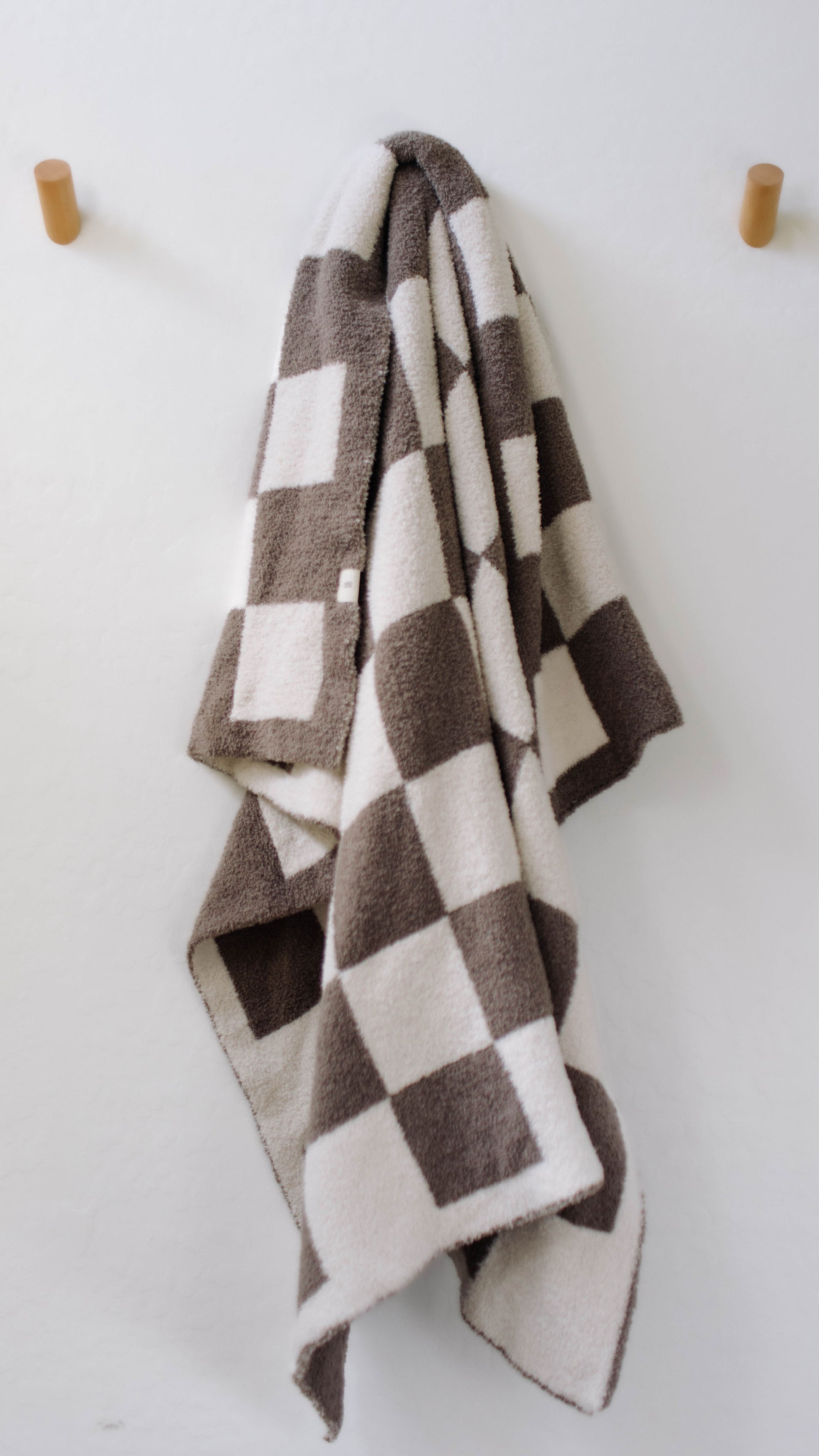 The Plush Blanket | Mushroom Checkerboard by forever french baby, a cozy creation made of microfiber feather yarn with a velvety-rich texture and adorned with a checkered pattern in shades of brown and white, hangs neatly on wooden hooks against a plain white wall.