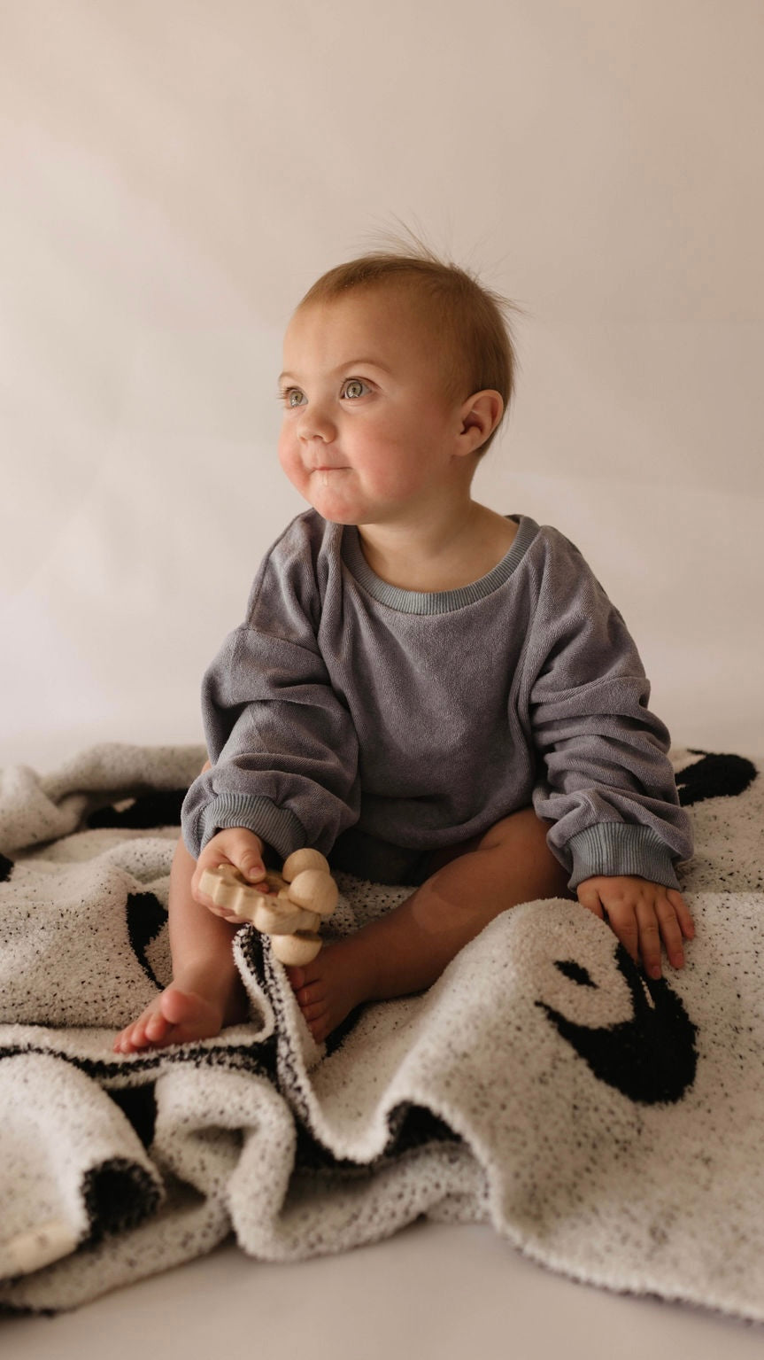 A baby with light hair sits on a soft, patterned "Plush Blanket | Black & White Yin Yang" made of microfiber feather yarn by forever french baby. The baby is wearing a loose-fitting gray sweater and holding a wooden toy in their left hand while looking slightly upward with a curious expression. The neutral and soft background enhances the serene atmosphere.