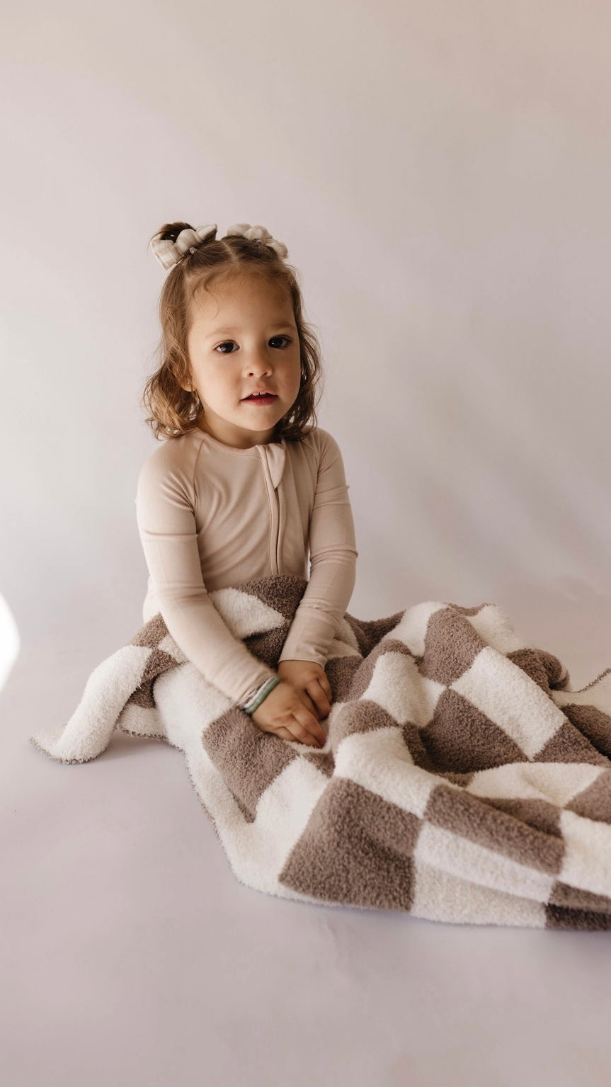 A young child with brown hair, pulled back with white bows, sits on the ground with a neutral expression. They are wearing a beige long-sleeve onesie and are partially wrapped in the Plush Blanket | Mushroom Checkerboard by forever french baby, featuring a large, fluffy design in shades of brown and white with a velvety-rich texture.