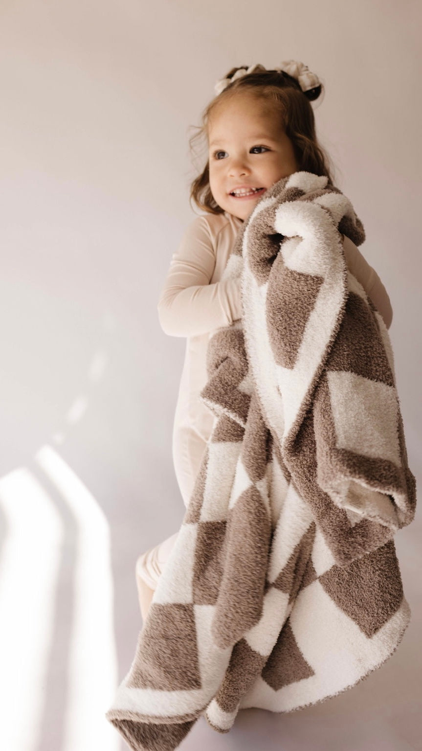 A young child with a cheerful expression holds the "Plush Blanket | Mushroom Checkerboard" from forever french baby, made from Oeko-tex certified materials. The child is dressed in light-colored clothing and has a bow in their hair. The background is neutral and softly lit.