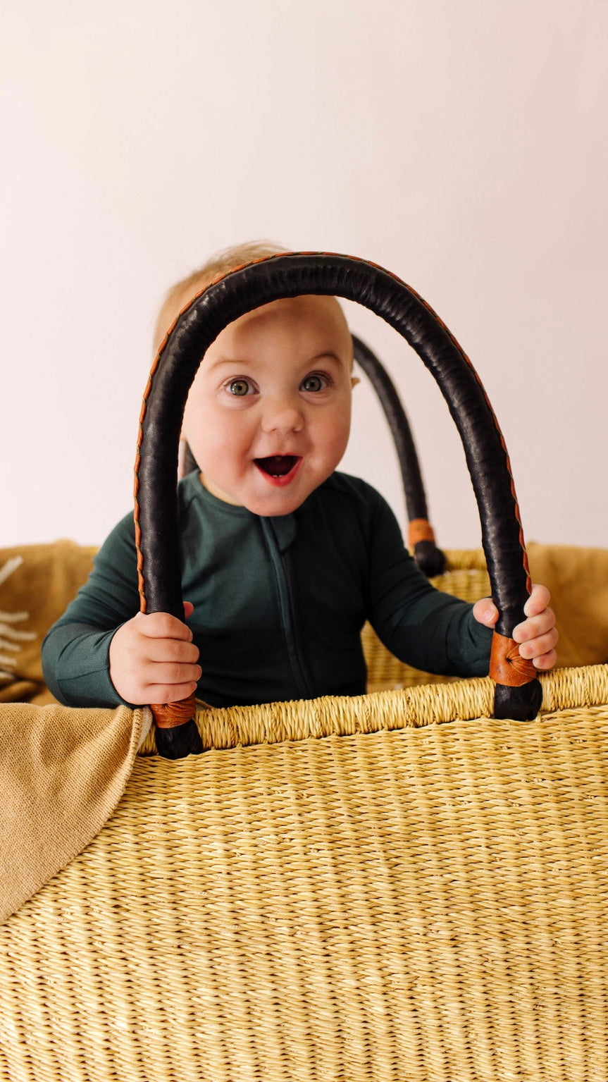 A smiling baby in Forever French Baby's Bamboo Zip Pajamas in Emerald stands inside a wicker basket, holding the black handles. A brown blanket is draped over one side of the basket against a plain light background.