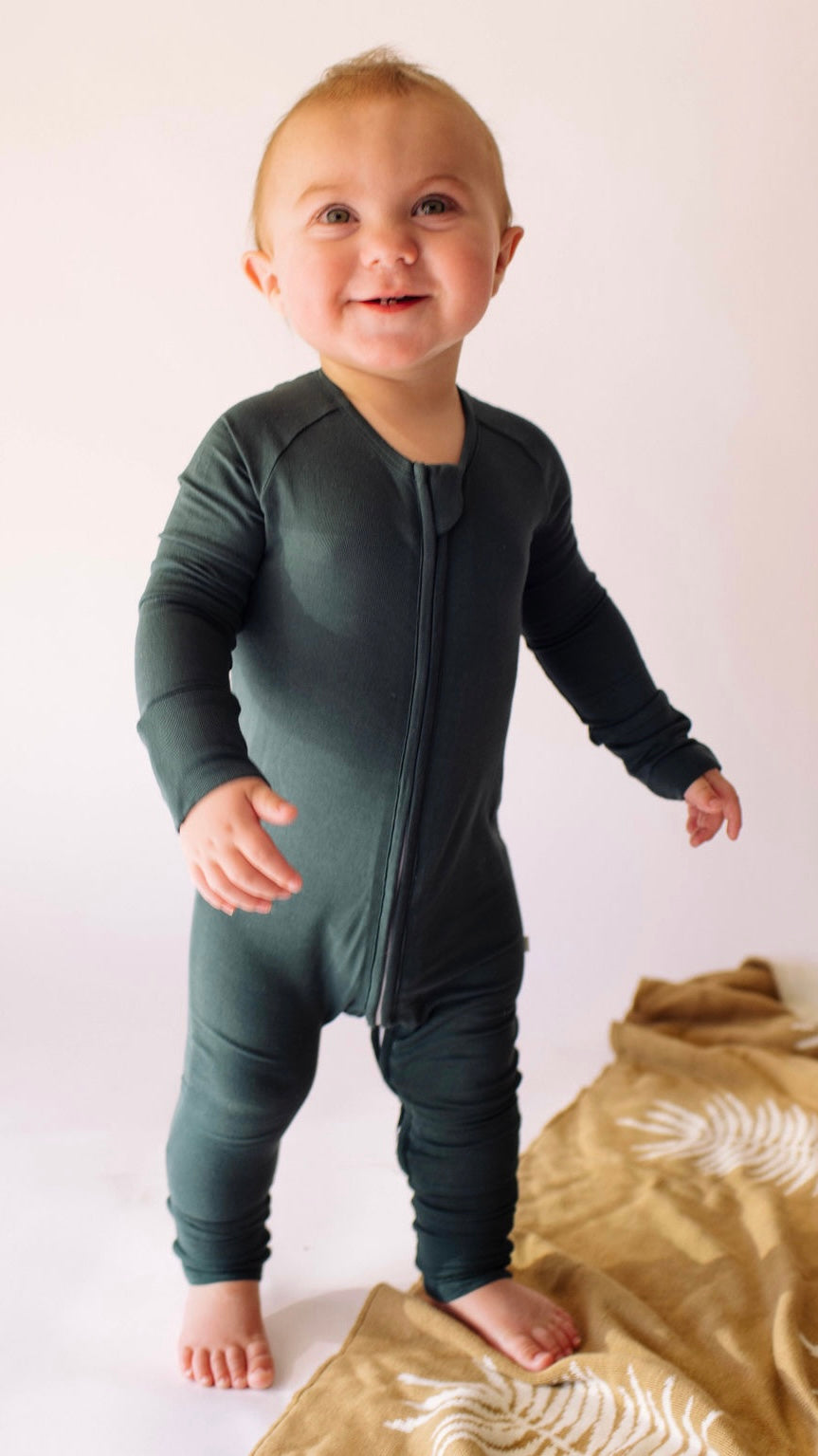 A smiling baby stands on a soft surface, wrapped in cozy hypoallergenic clothing. The Bamboo Zip Pajamas in Emerald by forever french baby ensure comfort with their front zipper, while a nearby brown blanket with white patterns adds a charming touch.