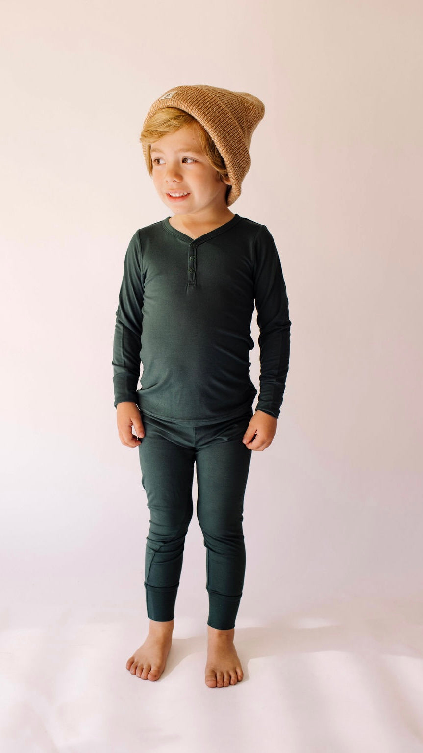 A child stands barefoot against a light background, smiling and looking to the side, while wearing "Emerald | Bamboo Two Piece Pajamas" by forever french baby and a brown knit beanie; this hypo-allergenic outfit promises year-round comfort.