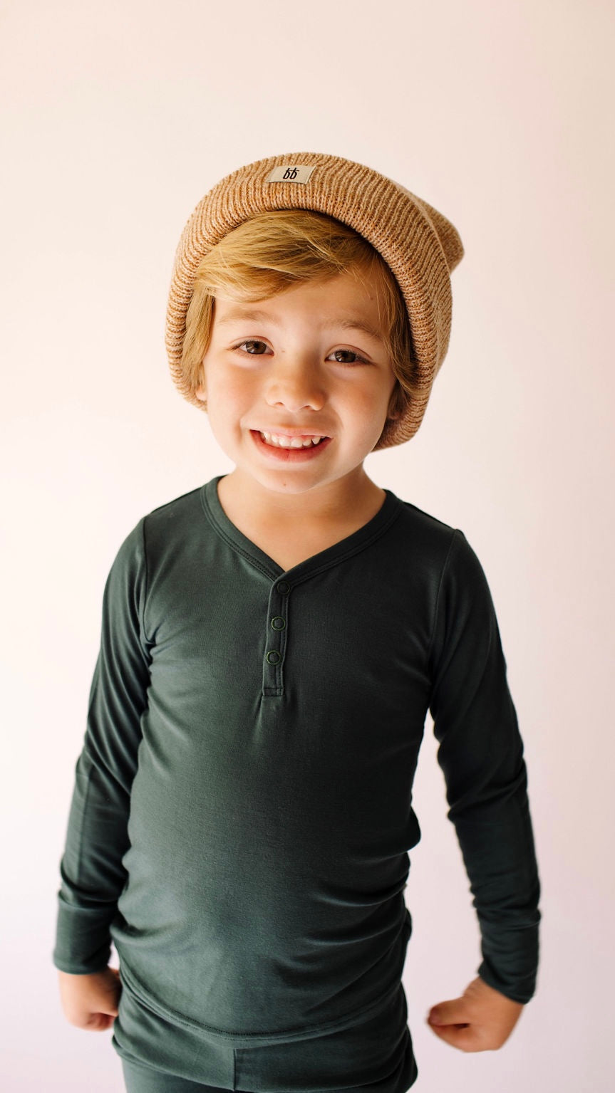 A young child smiles at the camera, wearing an Emerald Bamboo Two Piece Pajamas set by forever french baby and a tan beanie. The background is plain and light-colored.