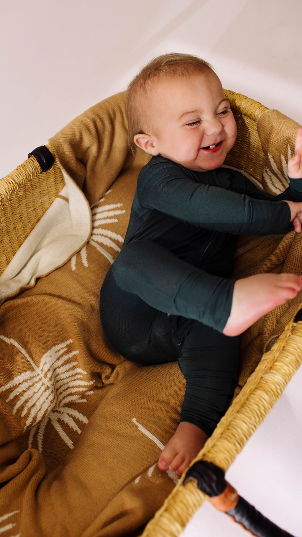 A happy baby, wearing forever french baby's Bamboo Zip Pajamas in Emerald, playfully lies in a wicker basket lined with a mustard-patterned blanket, their legs raised and one hand reaching for their foot.