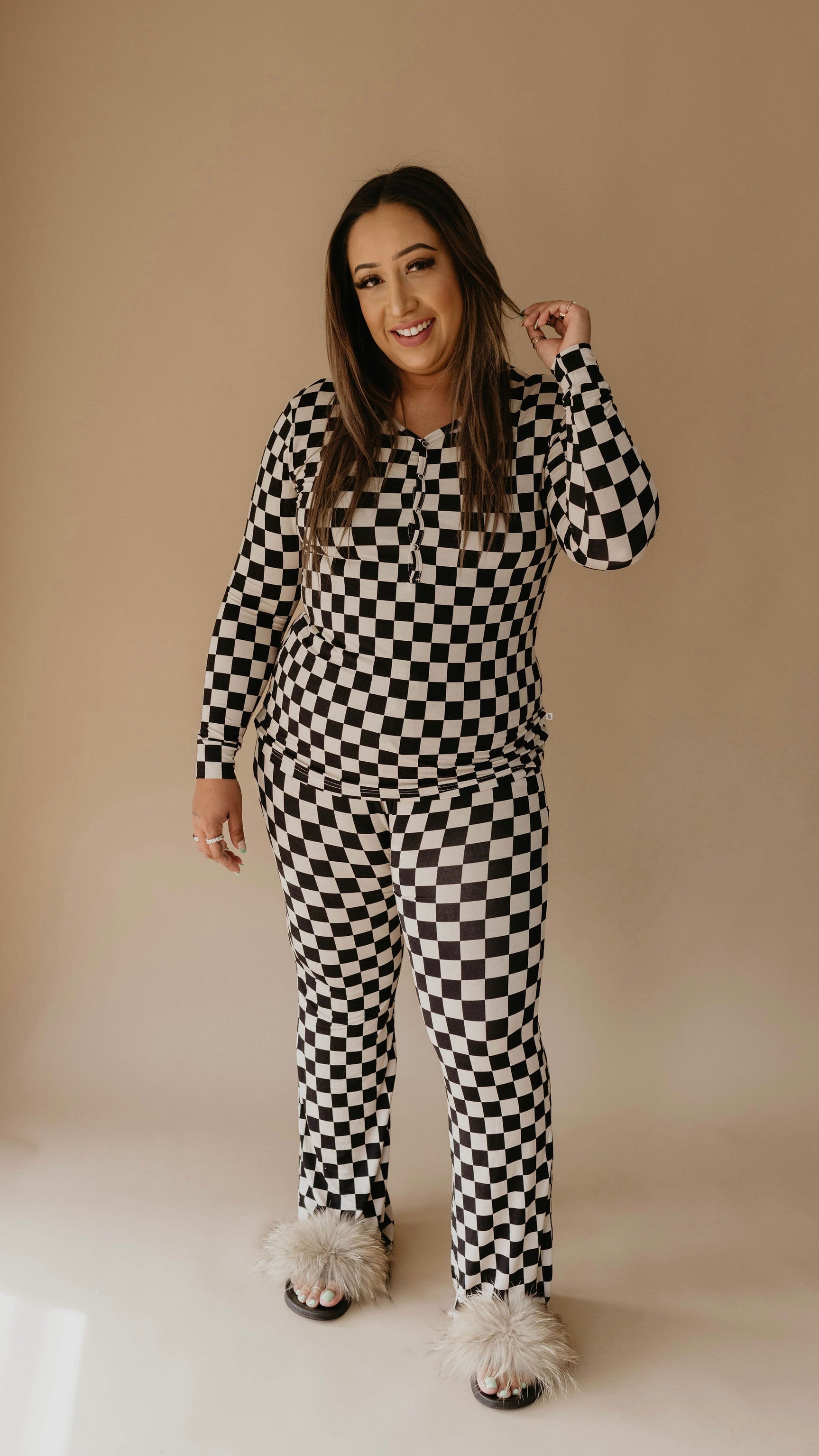 A person stands posing and smiling in forever french baby's Women's Bamboo Pajamas in Black Checkerboard, featuring a checkered black and white long-sleeved top and matching pants. They are wearing fluffy sandals and have long dark hair. The background is a plain, neutral color.