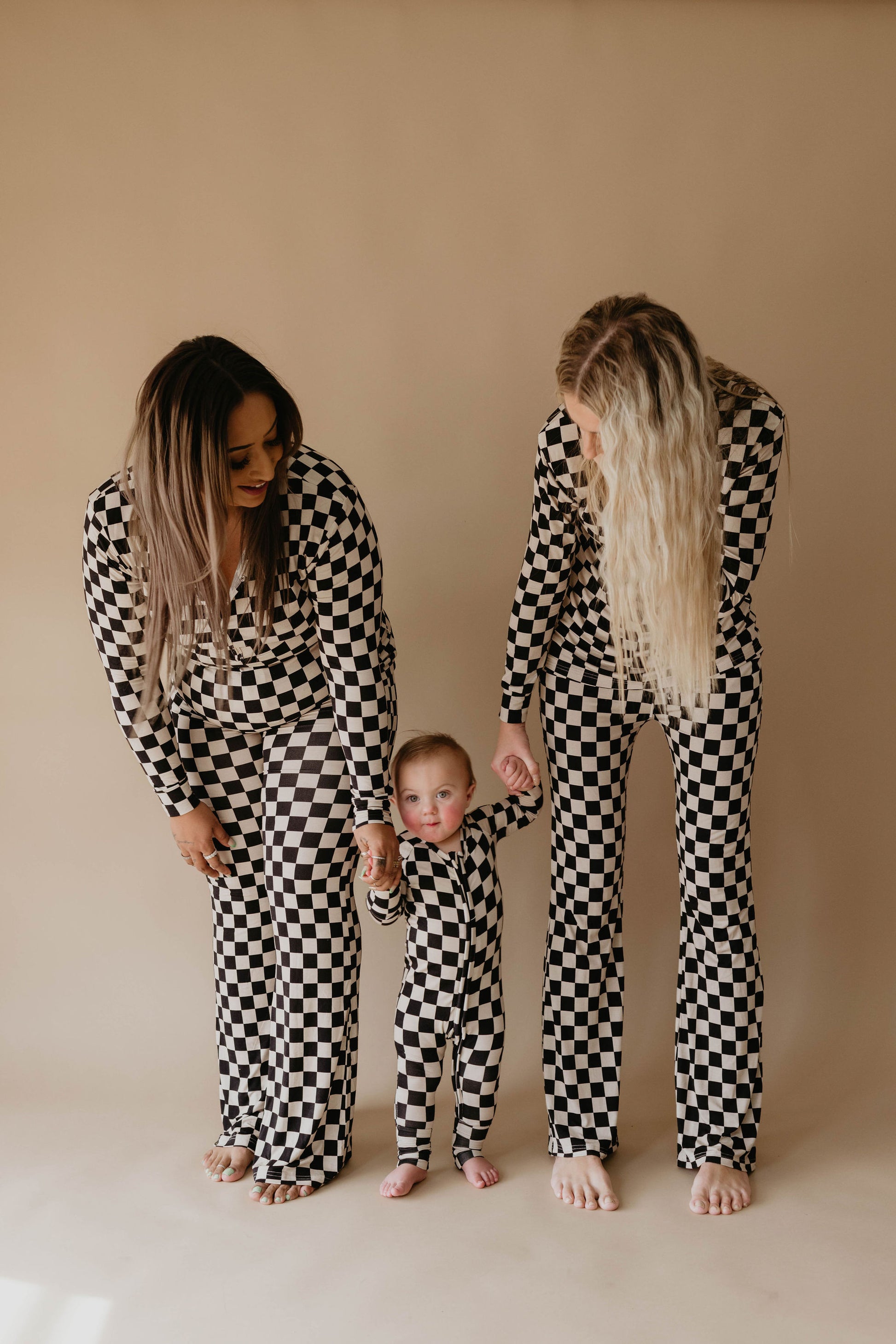 Two women and a baby stand together wearing matching black and white checkered outfits. The women are holding the baby's hands as they all smile down at the baby. They are dressed in stylish Women's Bamboo Pajamas in Black Checkerboard by forever french baby, standing against a plain beige background.