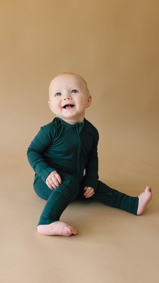 A smiling baby wearing forever french baby's Bamboo Zip Pajamas in Emerald sits barefoot on a light brown background, looking upwards with delight. The baby's light hair adds to the cheerful and serene scene.