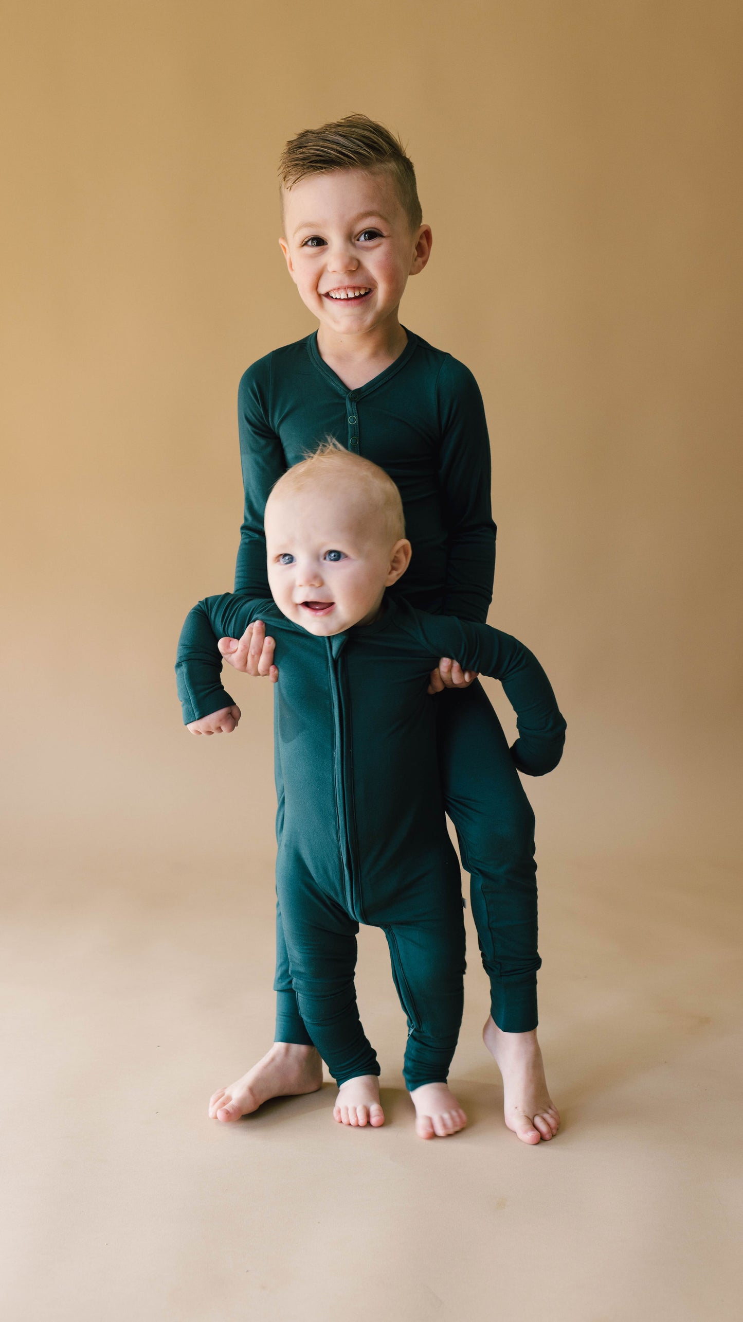 Two young children, both in matching Emerald Bamboo Zip Pajamas by Forever French Baby, are posed against a light brown background. The older child is standing, smiling and holding the baby from behind. Both children are barefoot and smiling toward the camera.