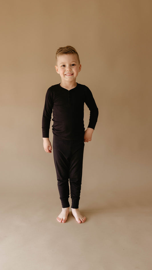 A young child stands barefoot on a beige floor against a matching beige background. The child has short hair and is wearing comfortable year-round, hypo-allergenic Bamboo Two Piece Pajamas in black, exclusively from forever french baby, with a slight smile on their face.