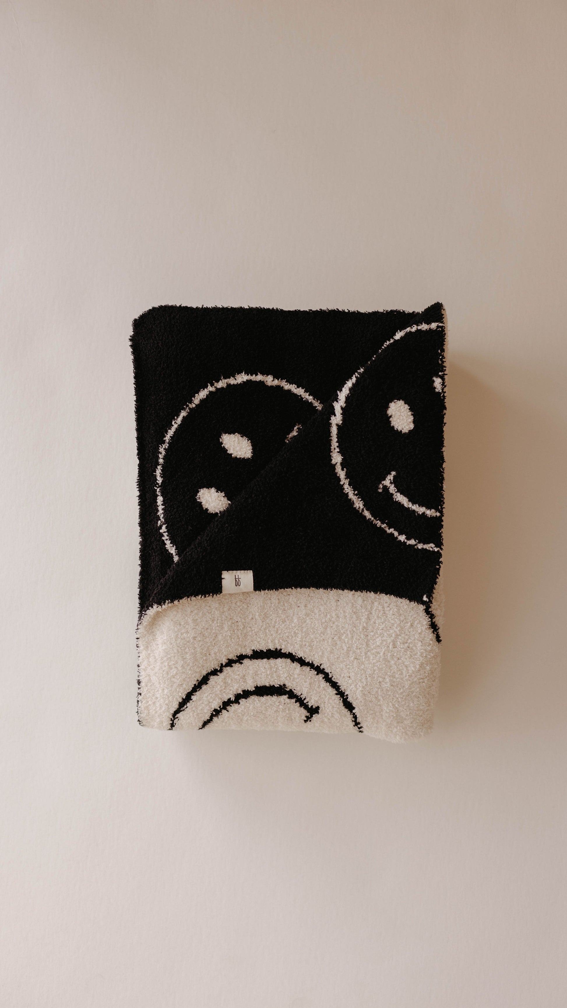 Introducing the "Plush Blanket | Just Smile Black & White" by forever french baby—a dreamy, soft blanket crafted from microfiber feather yarn. This luxurious black and white throw showcases a repeated smiley face design set against a beige background. One corner is turned over to reveal contrasting colors on each side, while a small white tag featuring the Oeko-tex logo is visible near the fold.