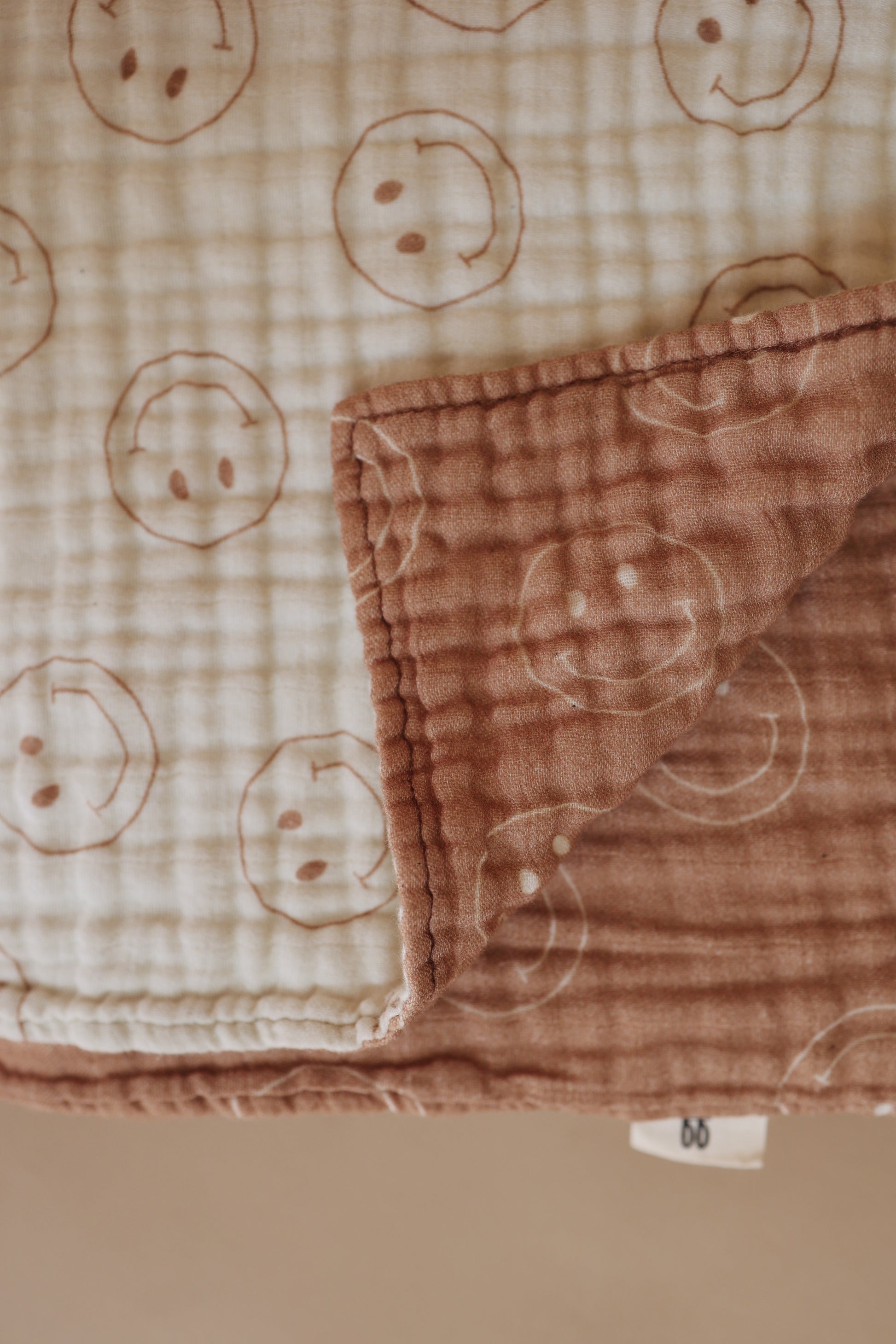 This Forever French Baby Muslin Quilt, named "Tan & Ivory Smile," is a folded, dual-colored reversible quilt. It features a beige side adorned with light brown smiley face patterns and a brown side with smiling faces in a lighter shade. The quilt boasts a textured, grid-like muslin cotton fabric design. A small label with "bb" is visible on one edge.