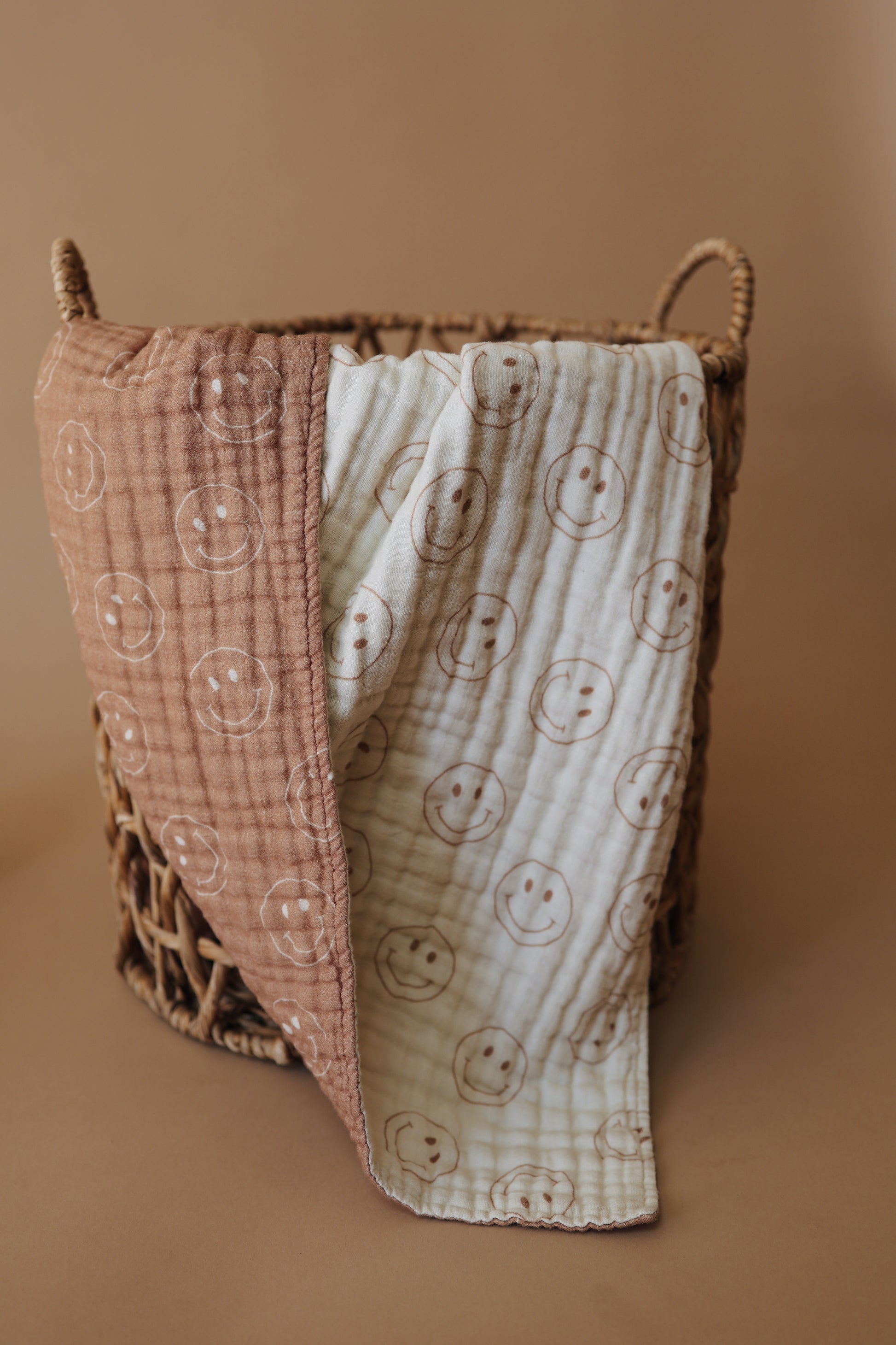 A woven basket contains a forever french baby Muslin Quilt | Tan & Ivory Smile draped over its side. The quilt features smiling face patterns on alternating tan and ivory sections, adding a touch of whimsy to the cozy scene. The background is a warm, neutral tone.