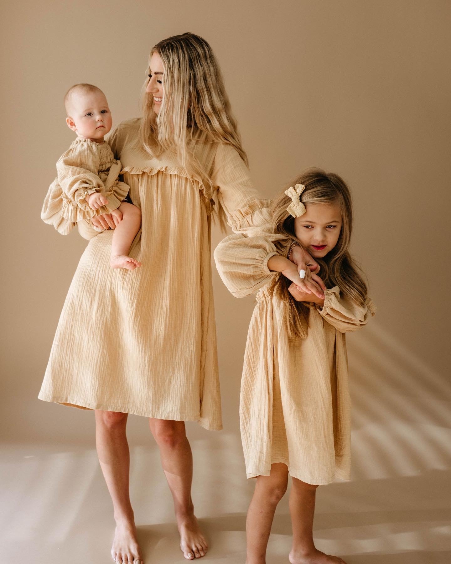 A woman stands barefoot holding a baby dressed in the "Golden Hour" ruffle romper from forever french baby, with a young girl beside her. All three wear matching beige dresses that cast a golden hue. The soft beige background creates a warm and harmonious scene.