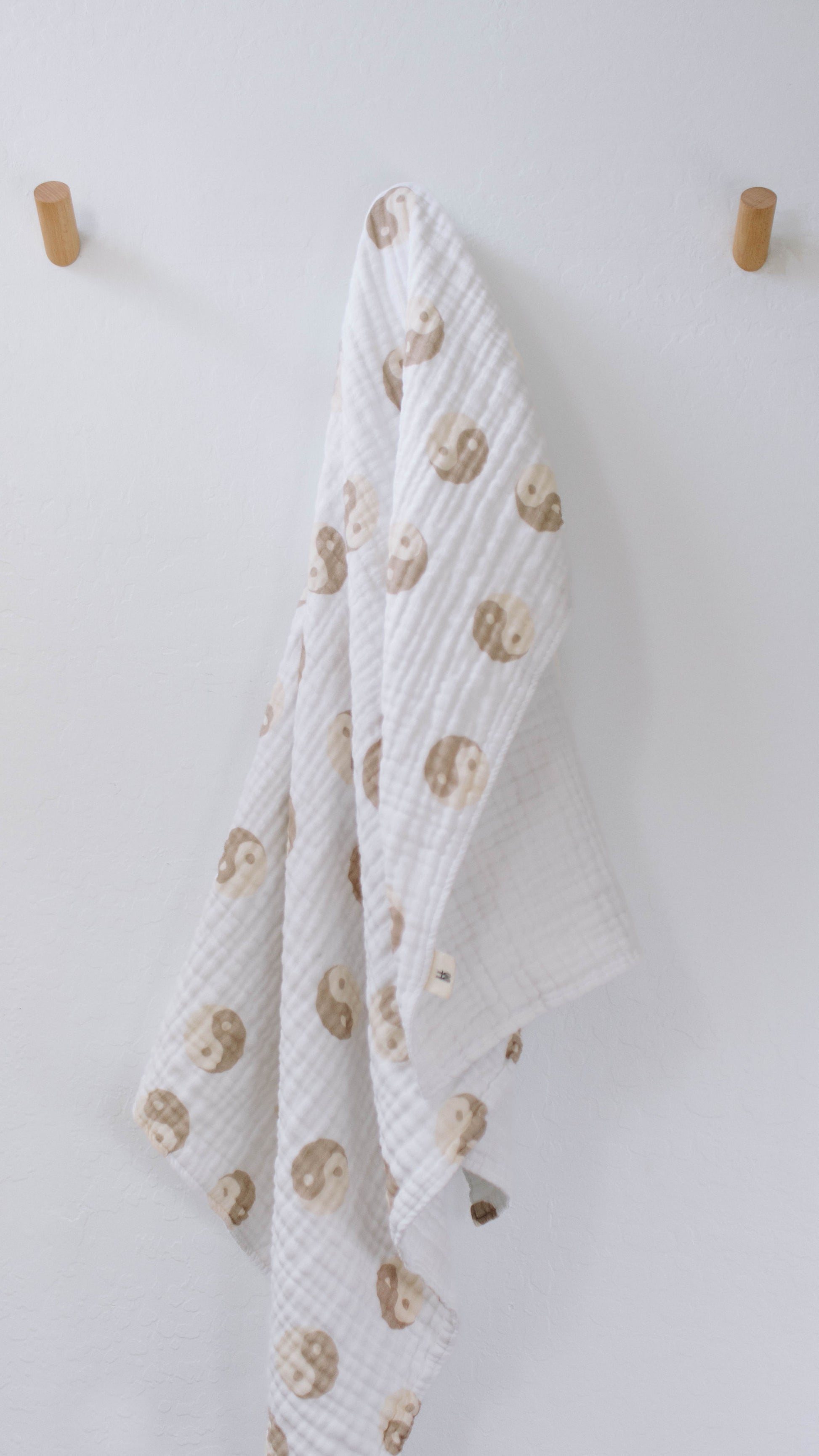 A 100% muslin cotton quilt adorned with light brown sloths hangs gracefully on a plain white wall between two wooden hooks. This charming Forever French Baby Yin Yang design brings a touch of whimsy and softness to any nursery.