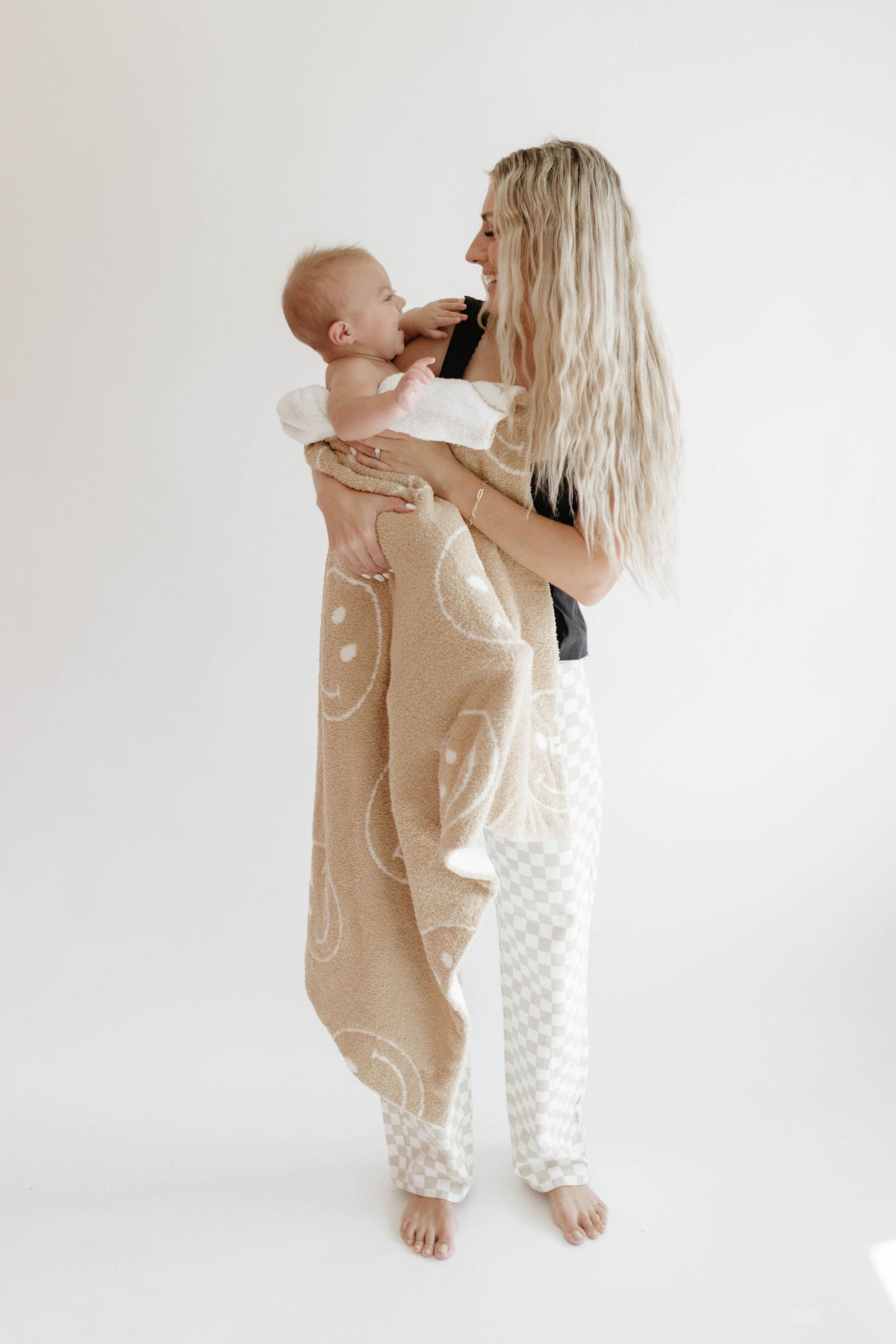 A barefoot woman with long, blonde hair is holding a smiling baby wrapped in the dreamy soft "Plush Blanket | Just Smile Black & White" by forever french baby. Both are dressed in casual, comfortable clothing and standing against a plain white background. The baby and woman are looking at each other, smiling.