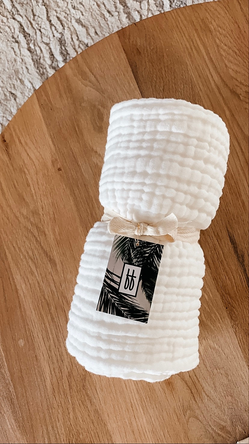 A neatly rolled, textured white crib sheet from Forever French Baby is tied with a beige ribbon and placed on a wooden surface. A tag featuring a palm tree design and the initials "bb" is attached to the ribbon. Remember to wash with like colors and tumble dry on low. A part of a textured white rug is visible in the background.