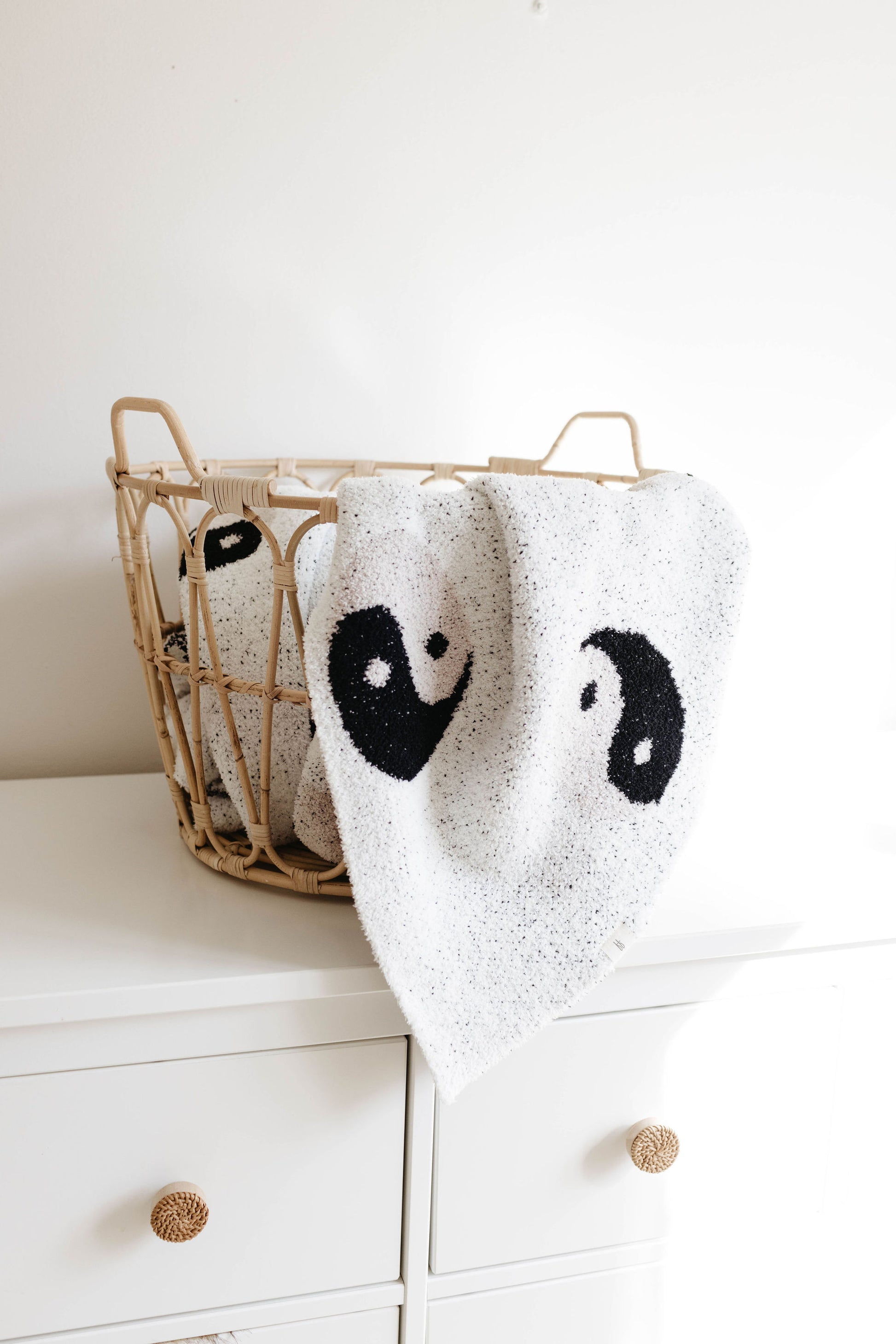 A white wicker basket sits on a white dresser. Inside the basket is a plush blanket by forever french baby, featuring a black and white yin-yang design. One corner of the blanket hangs over the basket’s edge, prominently displaying the intricate yin-yang symbol. The dresser has two drawers with round, knotted handles.