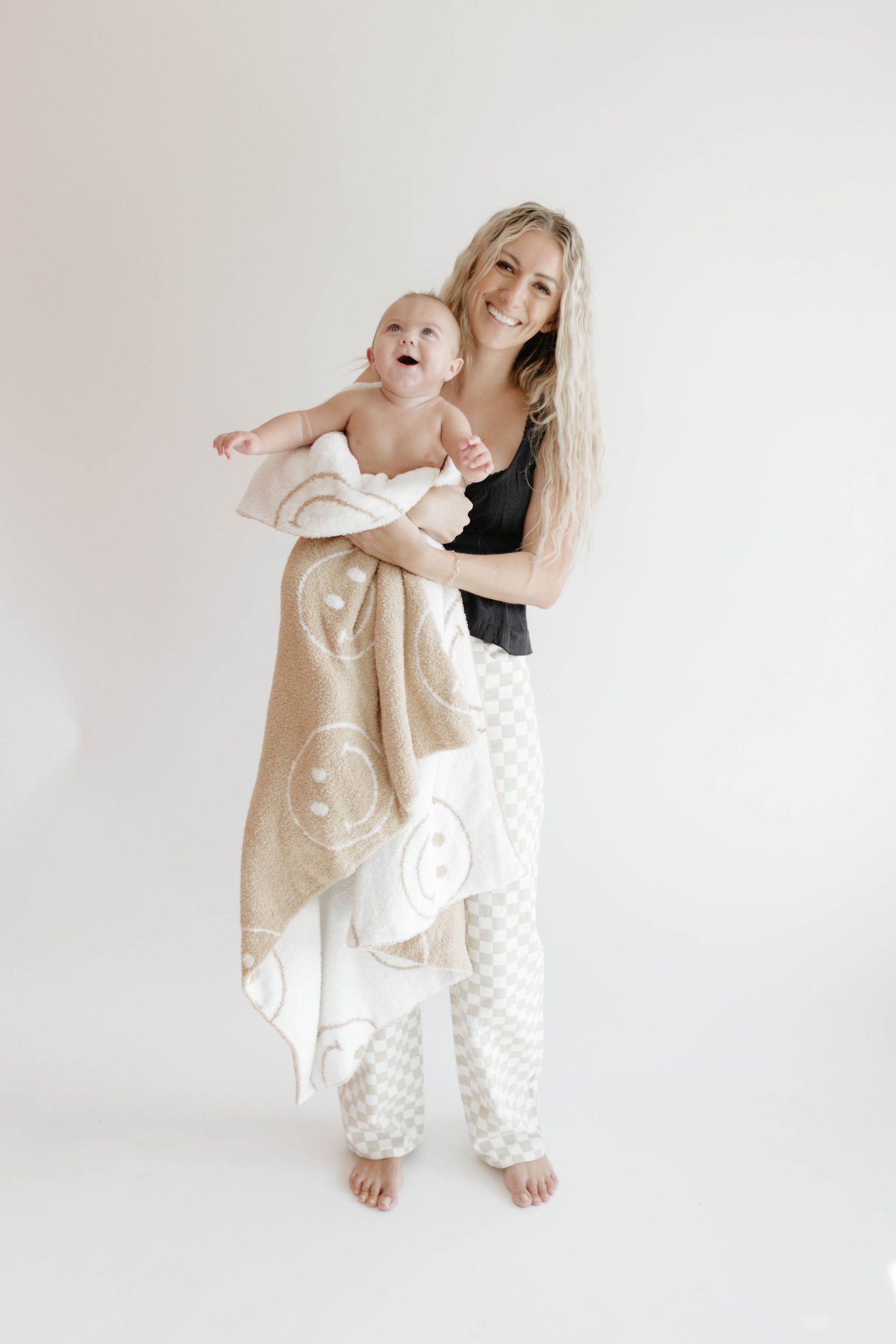 A woman with long blonde hair is holding a smiling baby wrapped in a Plush Blanket | Just Smile Black & White from the brand forever french baby. She is dressed in a black tank top and checkered pants, both barefoot, standing against a plain white background. Both appear happy and relaxed.