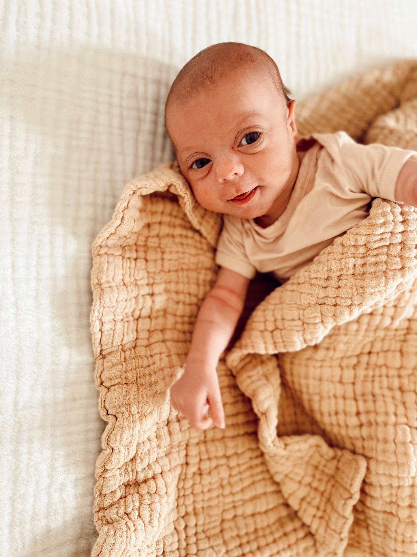 A baby with wide, curious eyes lies on a bed, partially wrapped in a textured beige newborn blanket. The baby, dressed in a light-colored onesie from Forever French Baby, has a slight smile and looks content. The background is softly complemented by the elegant Latte Quilt from forever french baby.